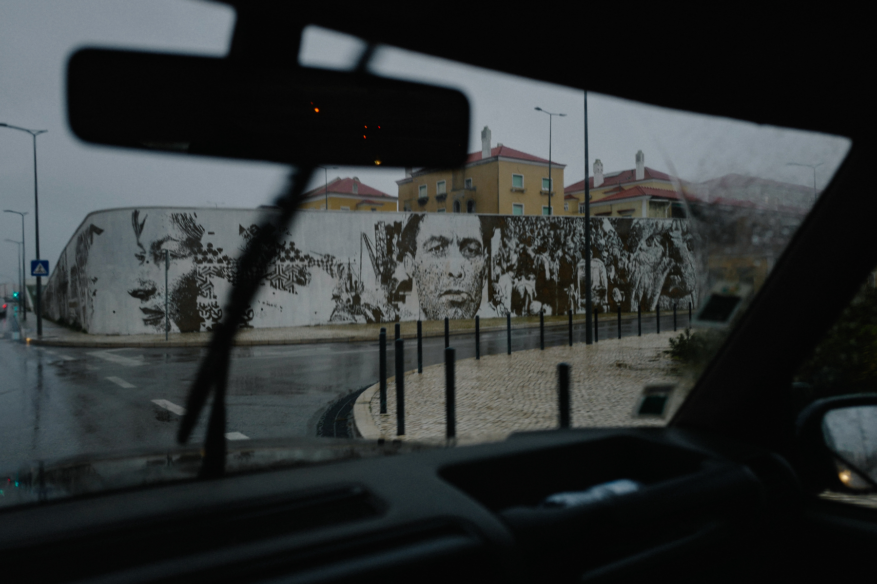 A view from inside a vehicle showing a windshield wiper and rearview mirror, with a street scene outside including a large mural on a curved wall, featuring stylized black and white faces and figures. The weather appears rainy.