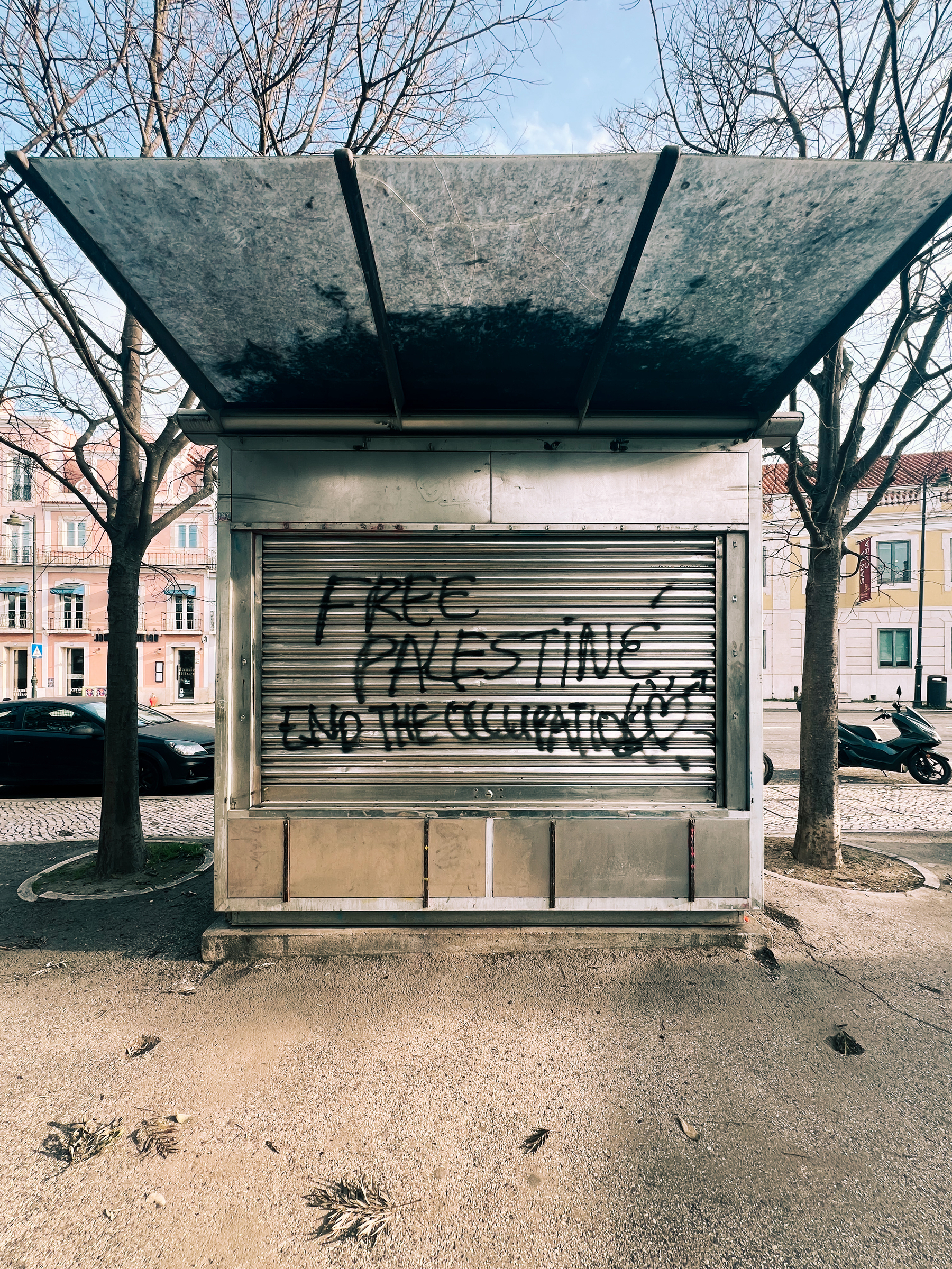 A shuttered kiosk with graffiti that reads &ldquo;Free Palestine End the Occupation&rdquo; on its metal closure, with trees and a clear sky in the background.