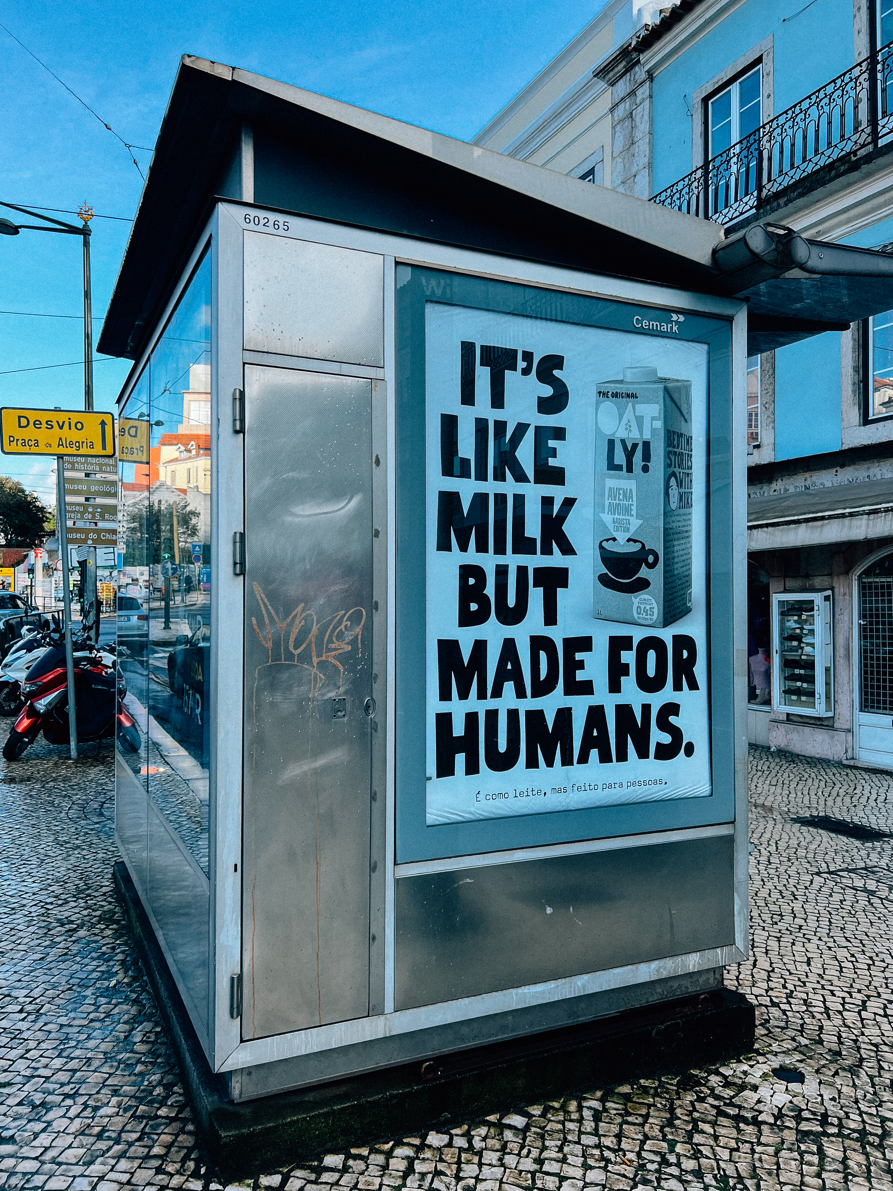 A kiosk featuring an advertisement for Oatly, a brand of oat milk, with the slogan &ldquo;It&rsquo;s like milk but made for humans.&rdquo; The shelter has graffiti on its side and is situated on a city street with cobblestones.