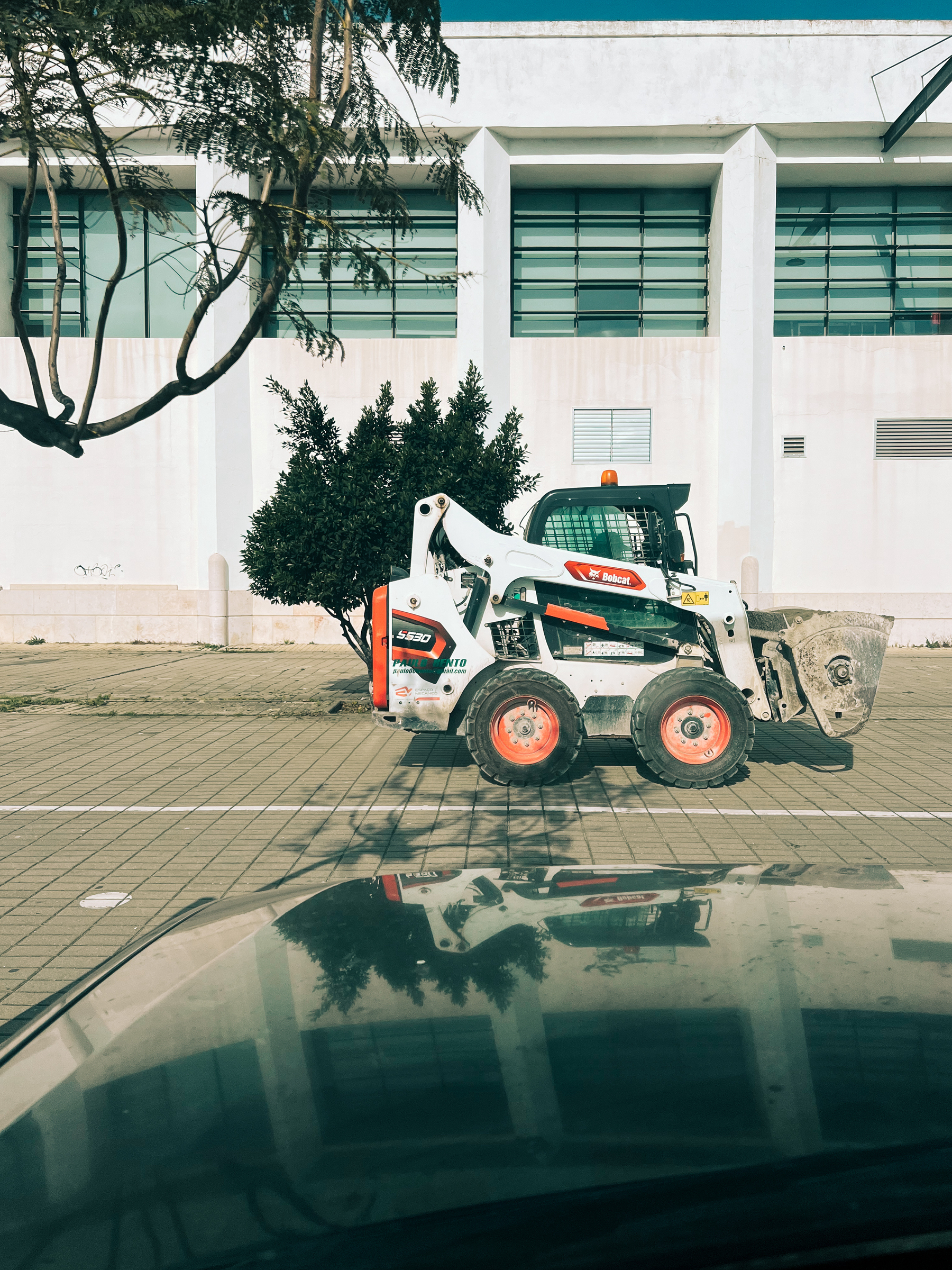 A skid-steer loader riding on a paved area with a modern building and trees in the background, the loader is reflected on the glossy hood of a car in the foreground.