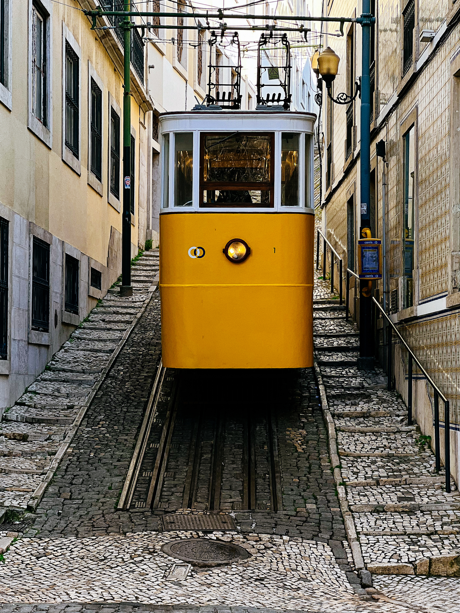 A yellow funicular tram moves up a steep, cobblestone street between traditional buildings, with power lines above and a streetlamp to the right.