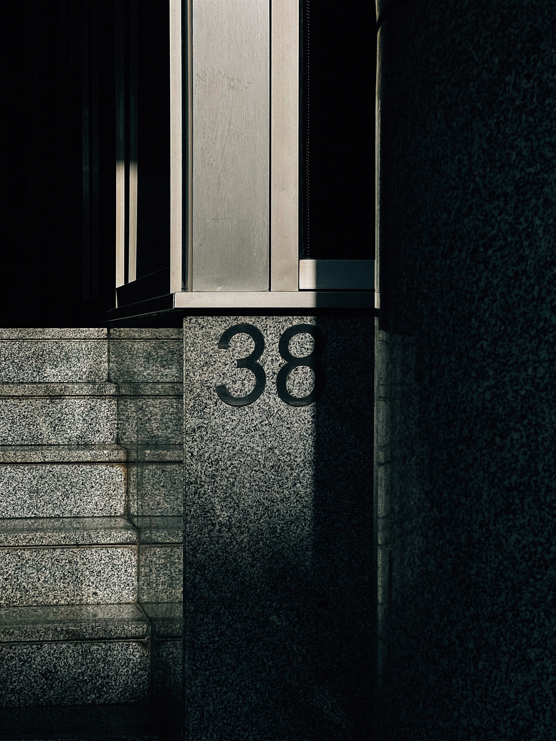 A number of 38 inscribed on a shadowed stone wall next to granite stairs and a column, with sunlight casting contrasting light and shadow effects.