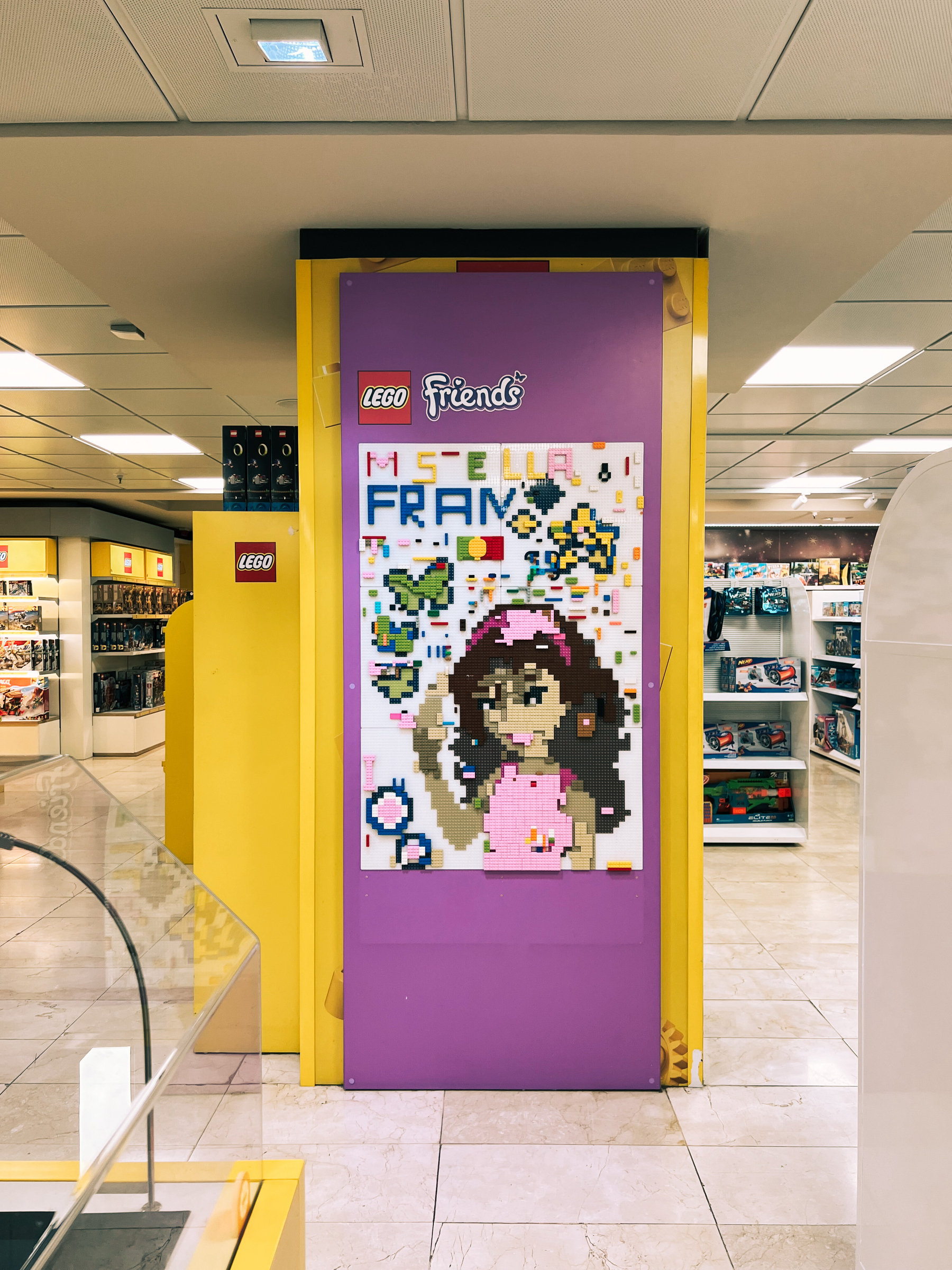 A colorful LEGO store entrance with a purple door featuring a LEGO Friends mosaic of a girl, and shelves with LEGO boxes in the background.