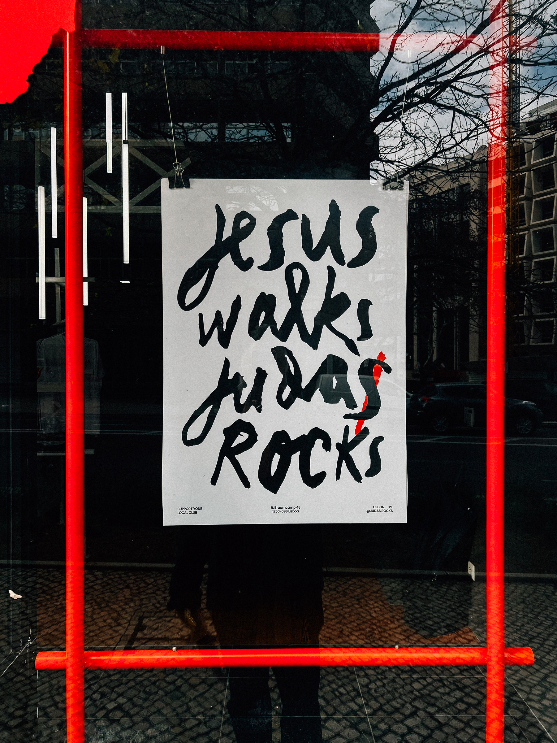 A poster with the phrases &ldquo;Jesus walks, Judas rocks&rdquo; displayed in a window, with a red frame and a reflection of a person standing in front of it.