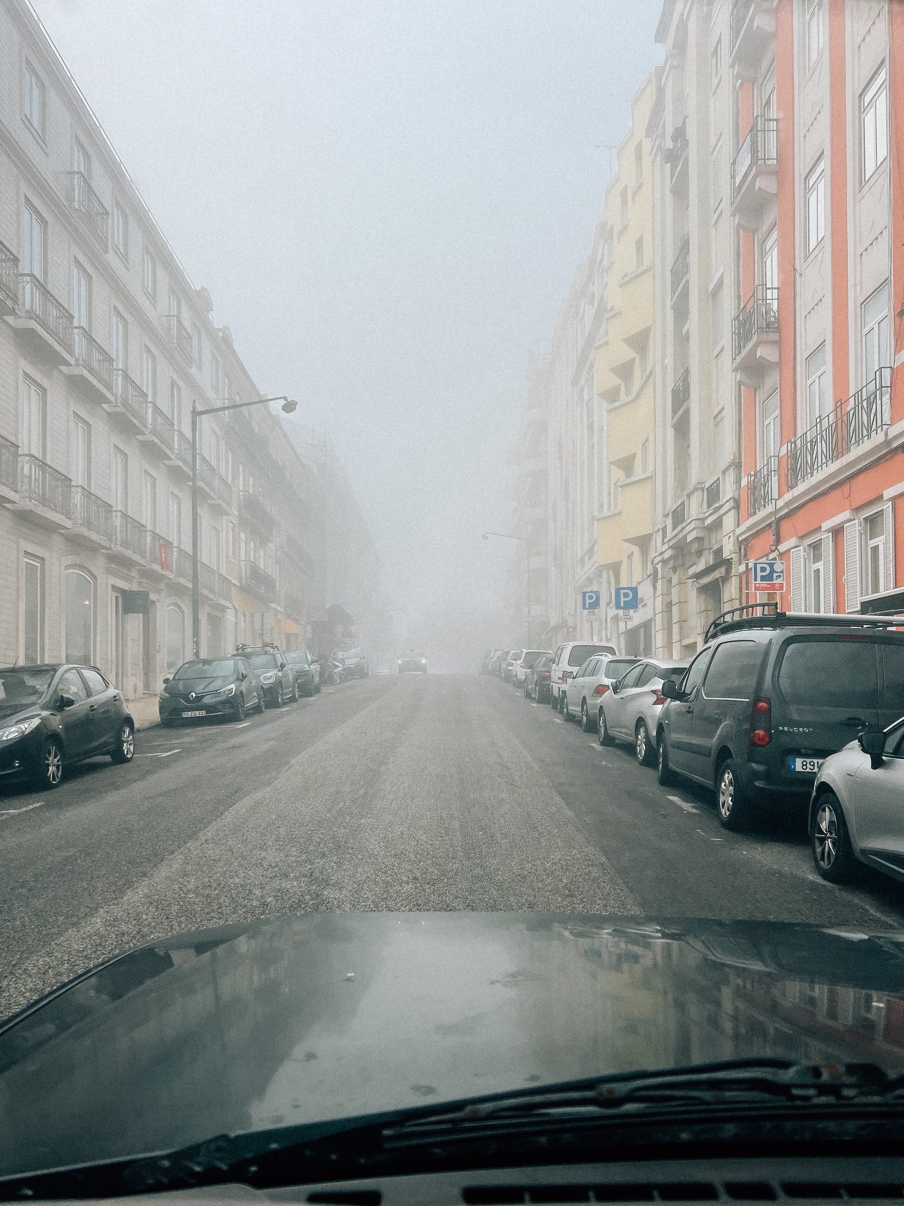 A view from inside a car showing a foggy street lined with parked cars and multistoried buildings, with visibility significantly reduced due to the dense fog.