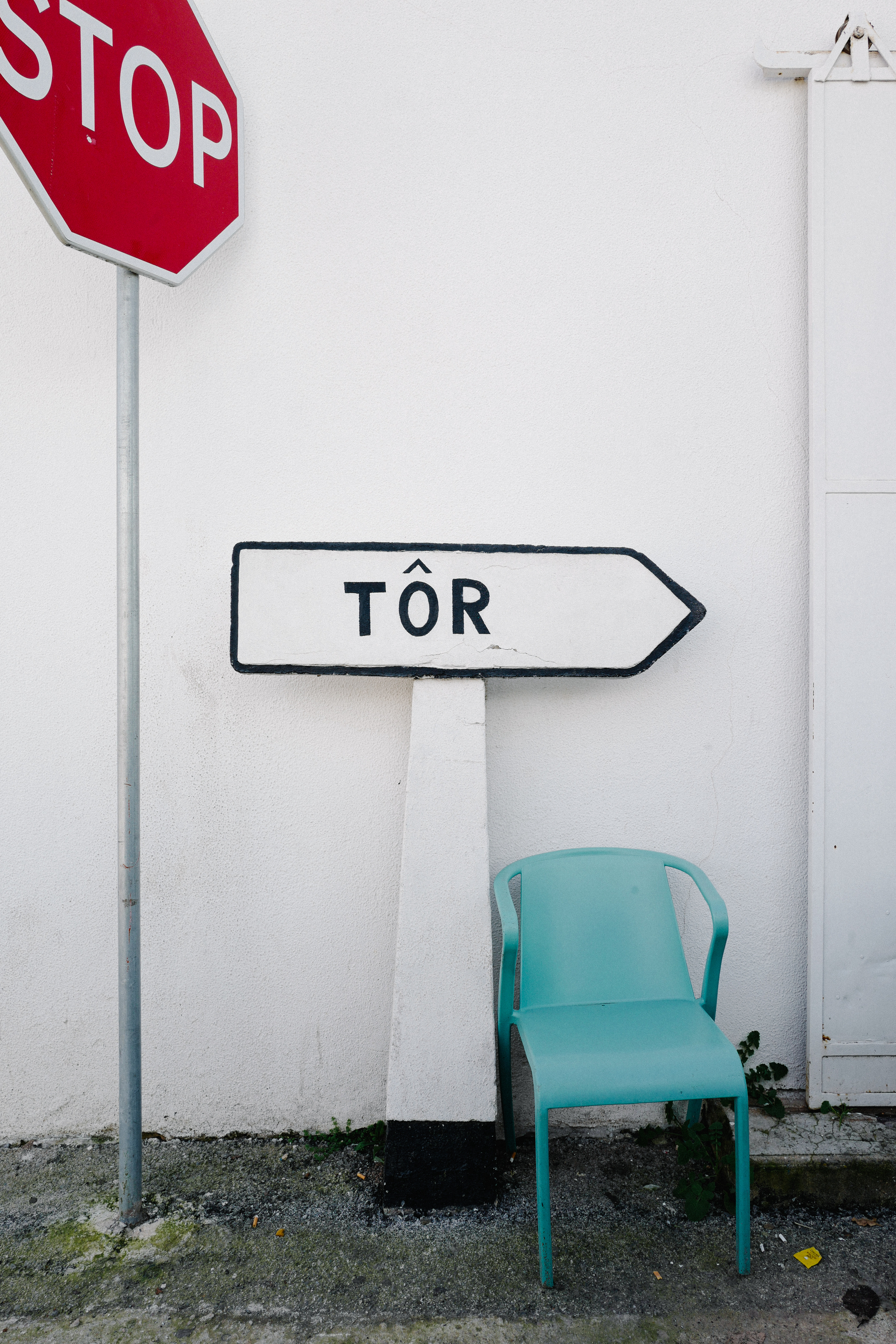 A stop sign, a directional arrow sign with the word &ldquo;TÔR,&rdquo; and a single turquoise chair against a white wall.