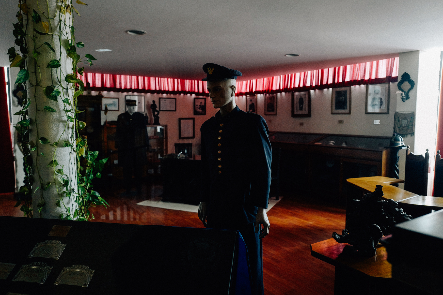 A dimly lit room displaying firemen memorabilia, with a mannequin dressed in a formal uniform at the center, flanked by showcases and framed items on the walls.