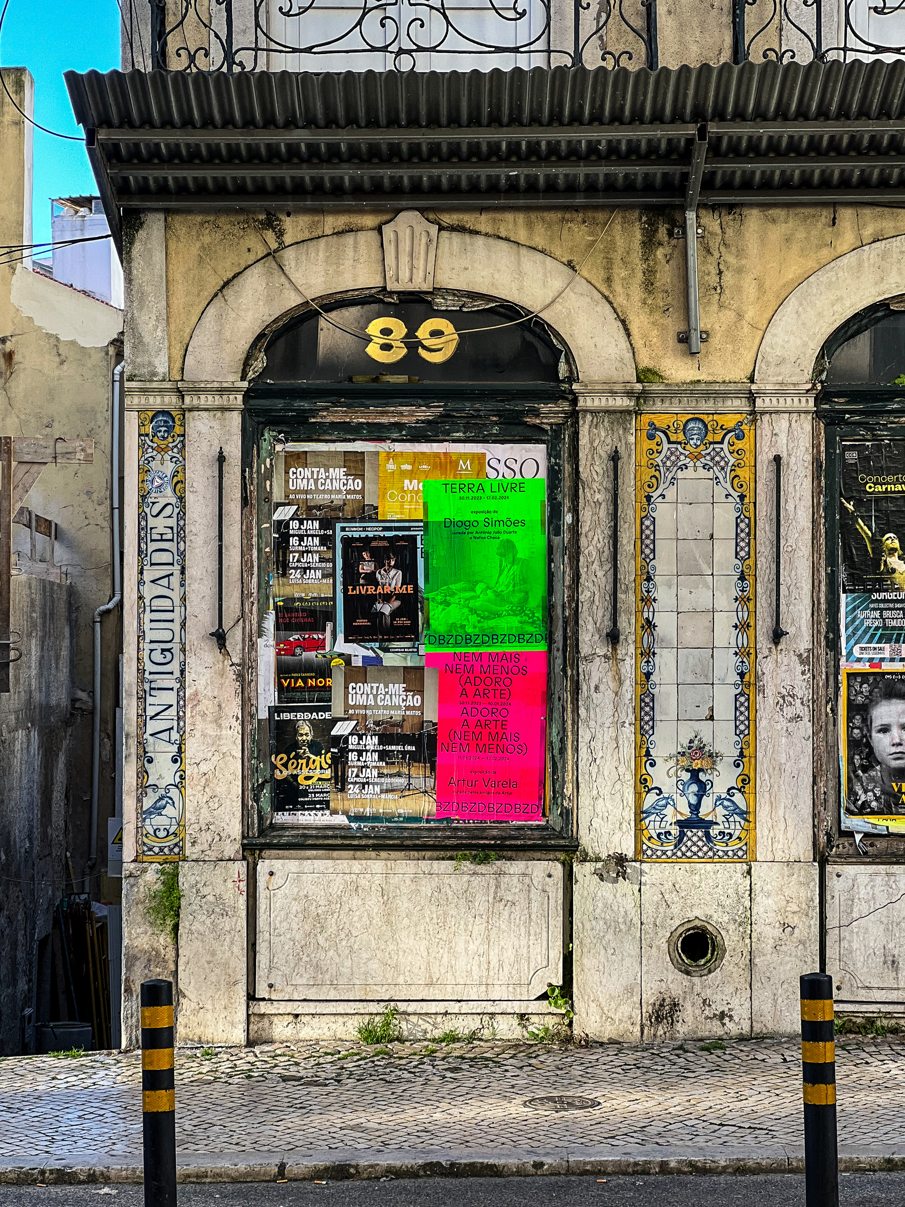An old storefront with faded paint and decorative tiles, displaying numerous colorful event posters and a sign with &ldquo;89&rdquo; above the window.