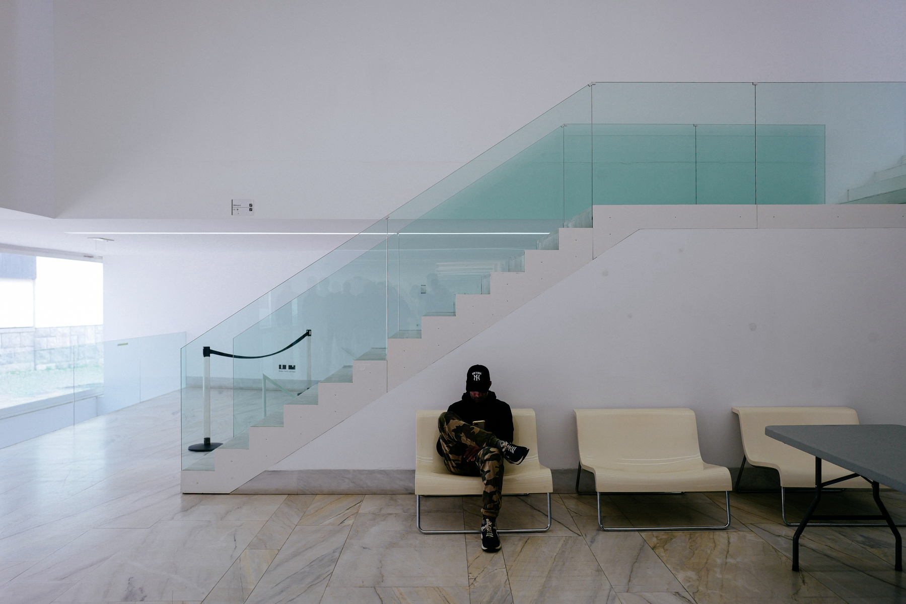 An individual wearing a cap and camouflage pants is seated on a chair in a modern interior with a glass-paneled staircase, white walls, and marble flooring. There are two empty chairs and a table beside them.