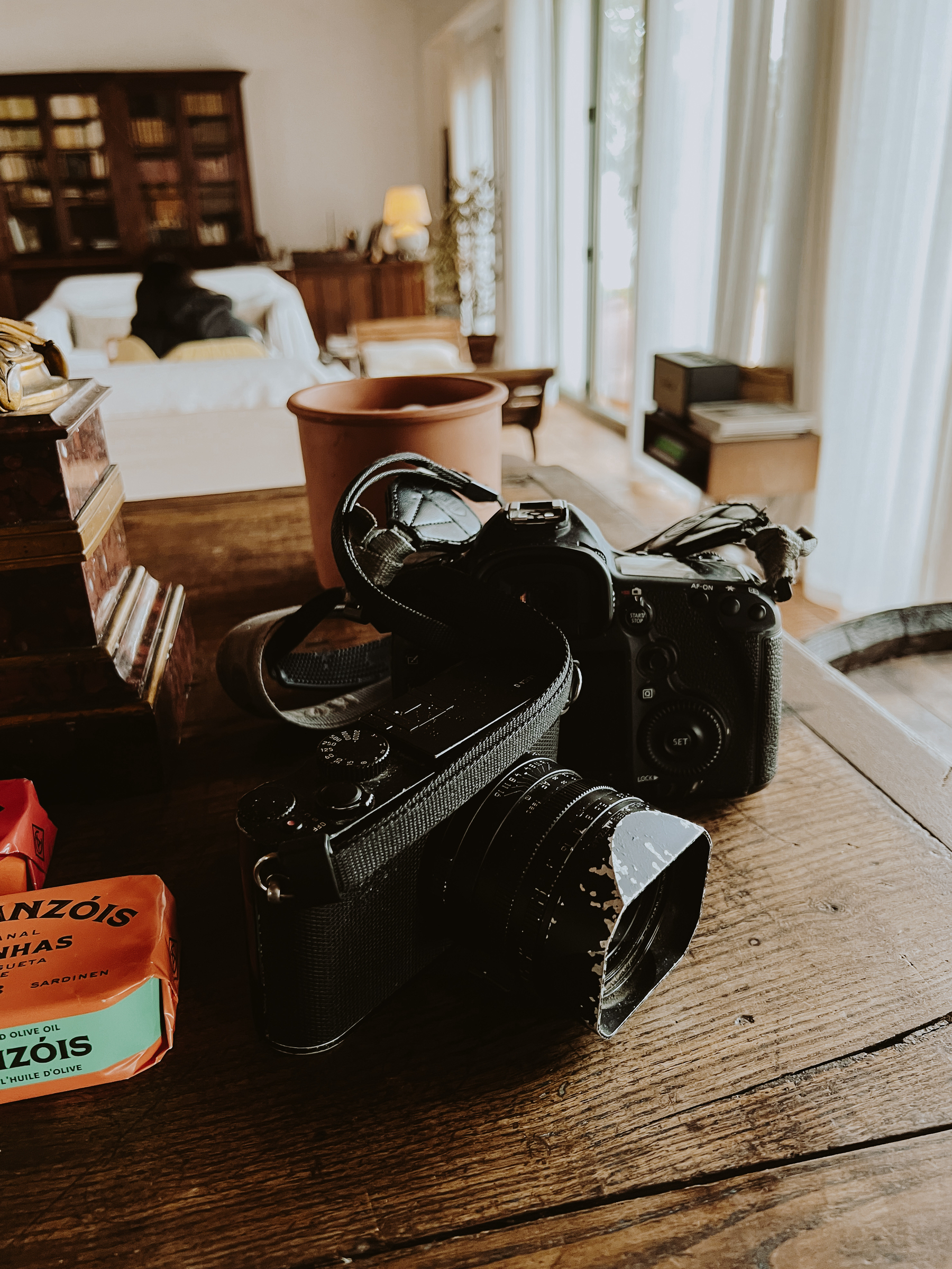 A camera with a strap placed on a wooden table, with its lens hood visibly worn and chipped. In the background, there&rsquo;s a cozy room with bookshelves, a bed, and a lamp, all bathed in warm light.
