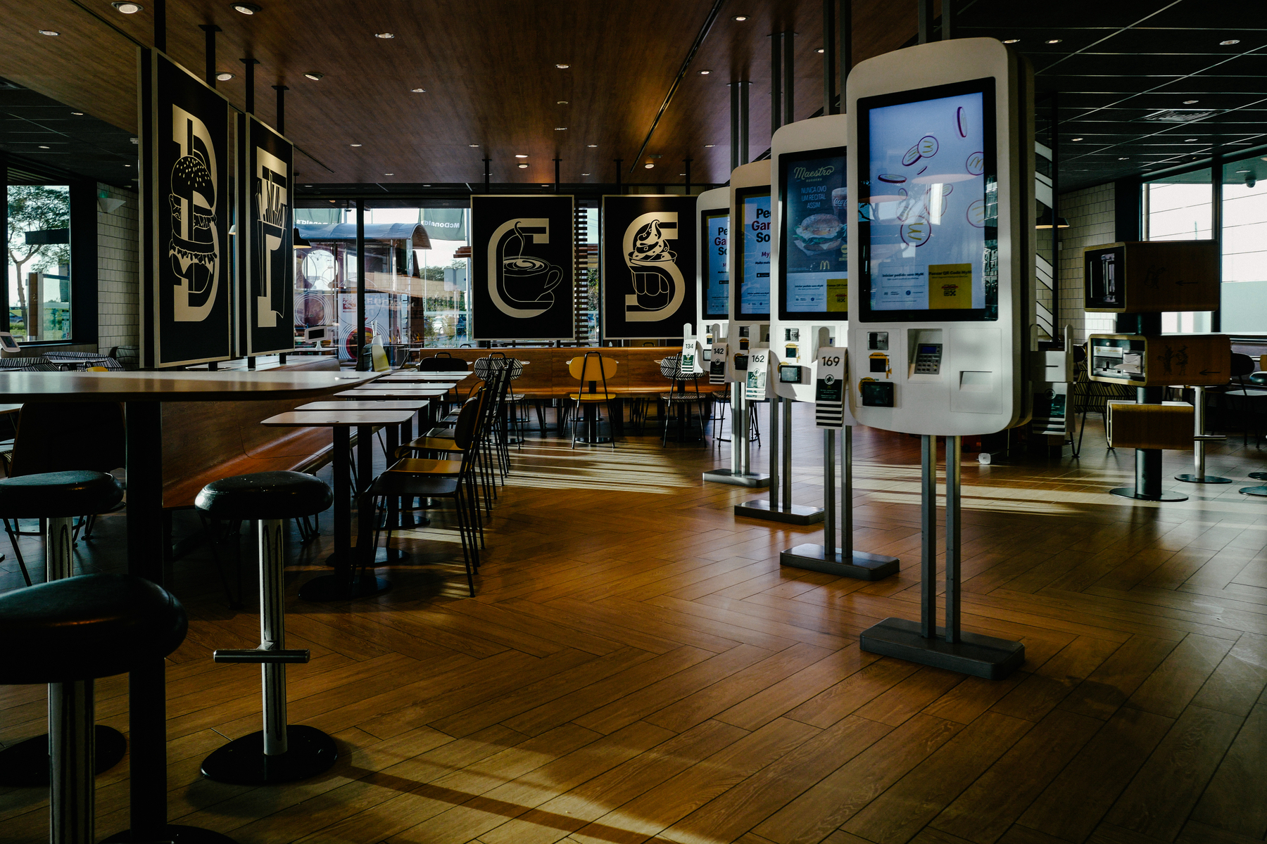 Interior of a modern fast food restaurant with digital ordering kiosks, assorted dining tables and chairs, and large graphic art on the walls.