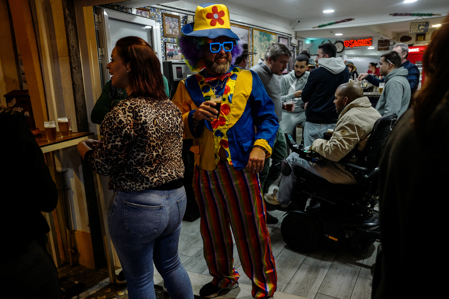 A person in a colorful clown costume with a hat, sunglasses, and wig stands with a drink in hand in a crowded indoor space, with other people visible in the background.