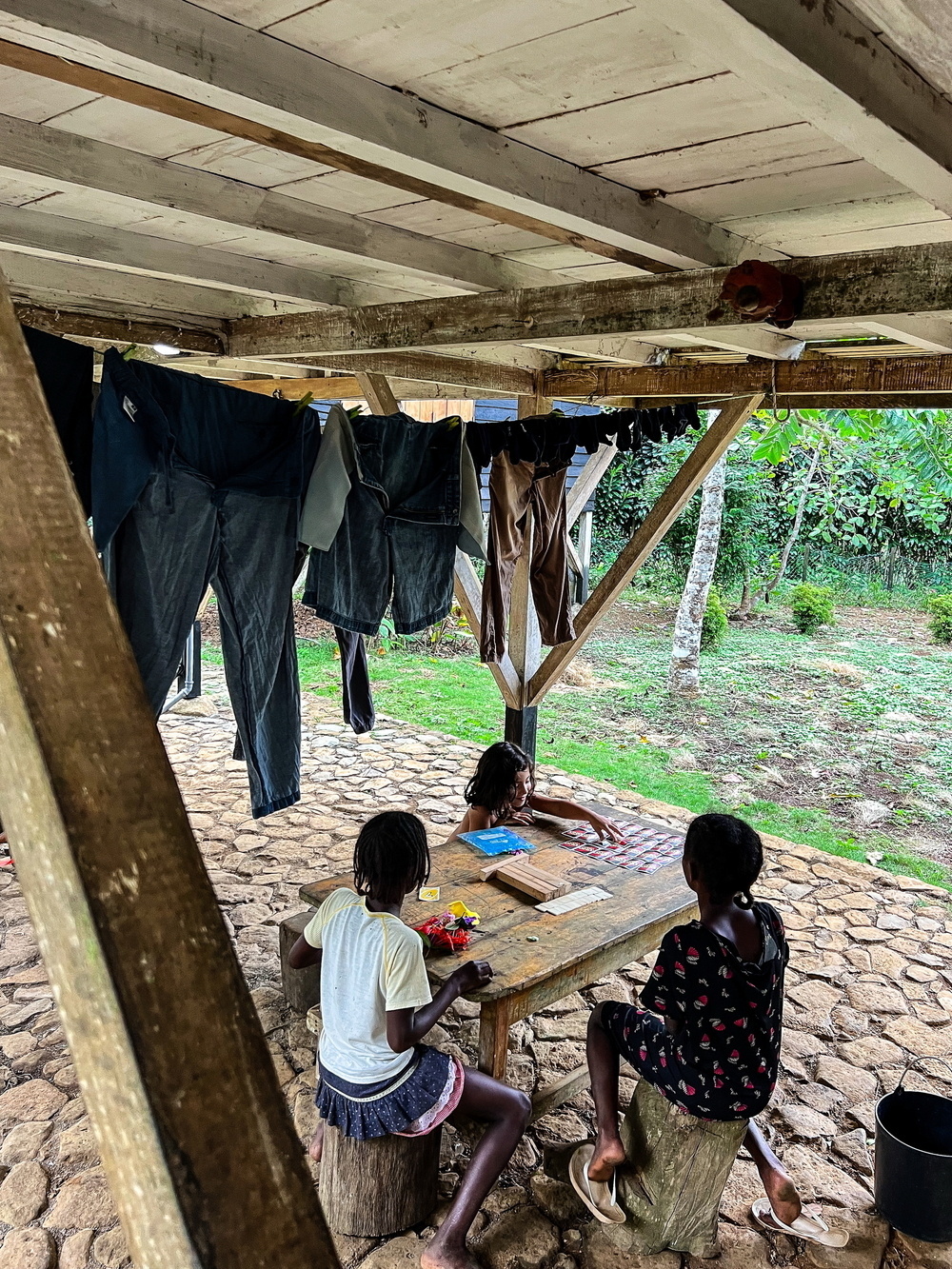 Children sit at a wooden table under a rustic shelter with a stone floor. Clothes hang on a line above them.