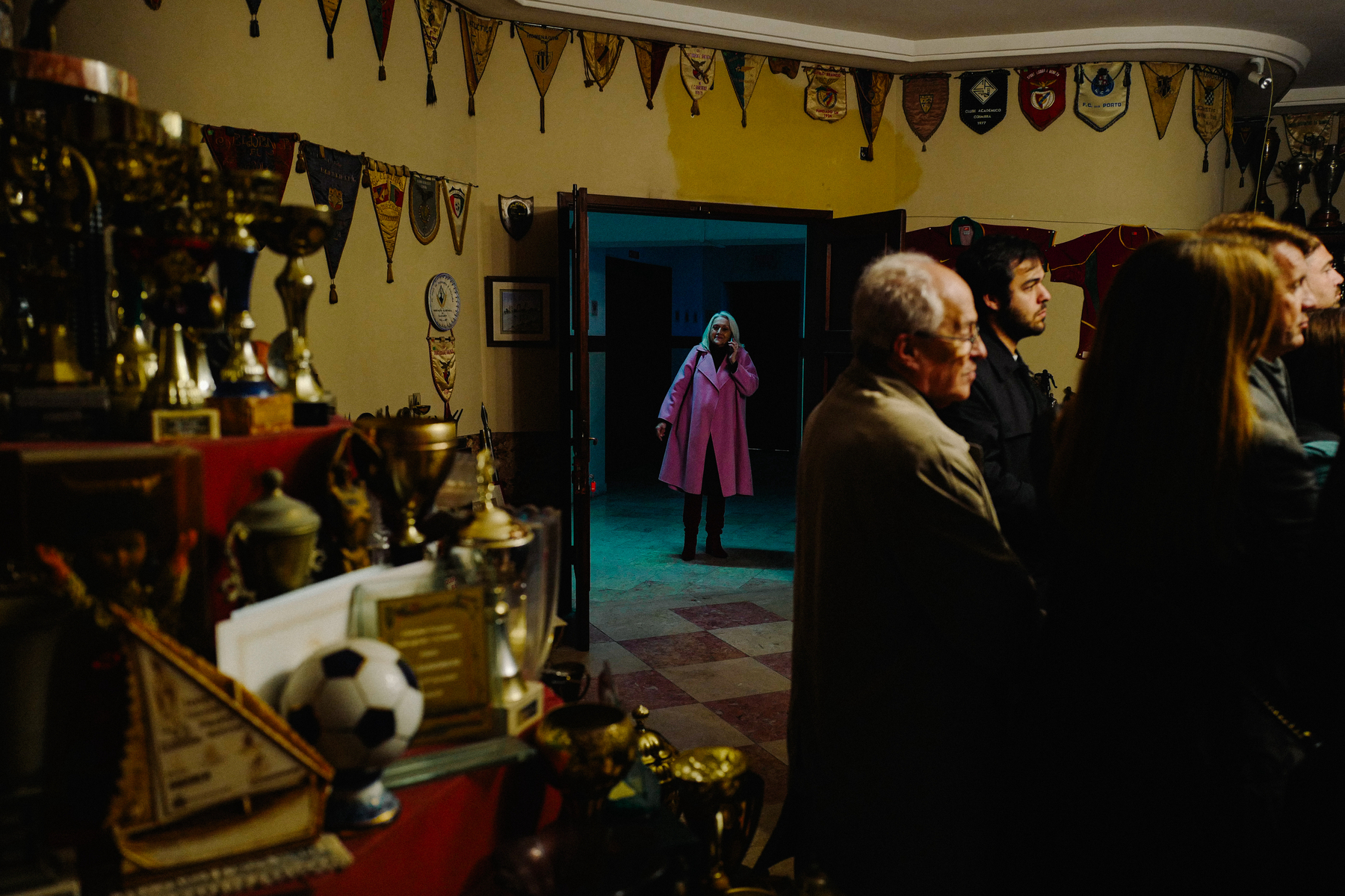 A trophy room filled with various awards and pennants with people gathered. In the doorway stands a person in a pink coat.