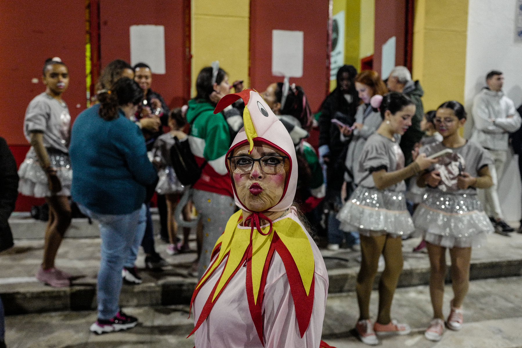 A person dressed in a colorful chicken costume with face paint stands in the foreground among a crowd of people, some of whom are wearing matching outfits and using their phones. The background is blurry, emphasizing the individual in the chicken costume.