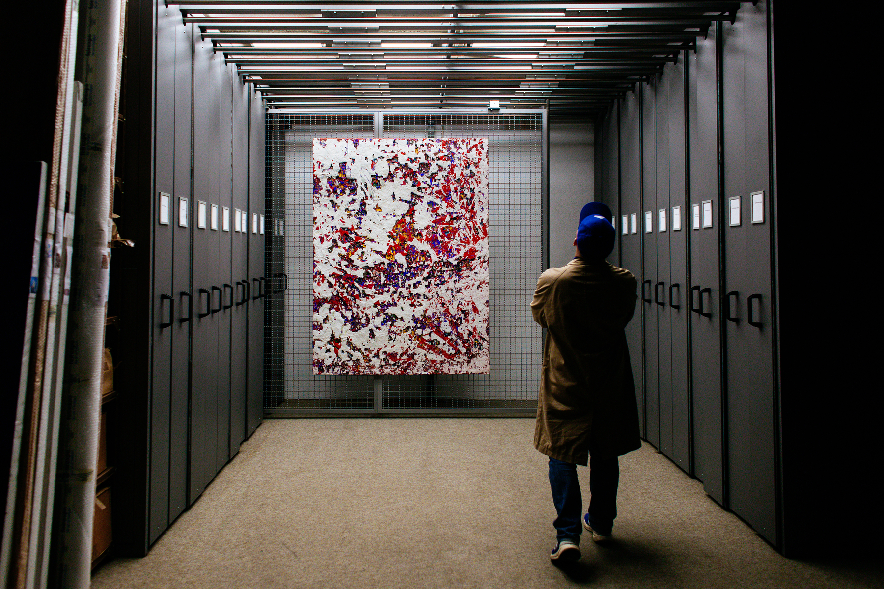 Person in a blue cap and beige coat observing a large abstract painting displayed on a wire mesh wall at the end of a corridor lined with gray lockers.