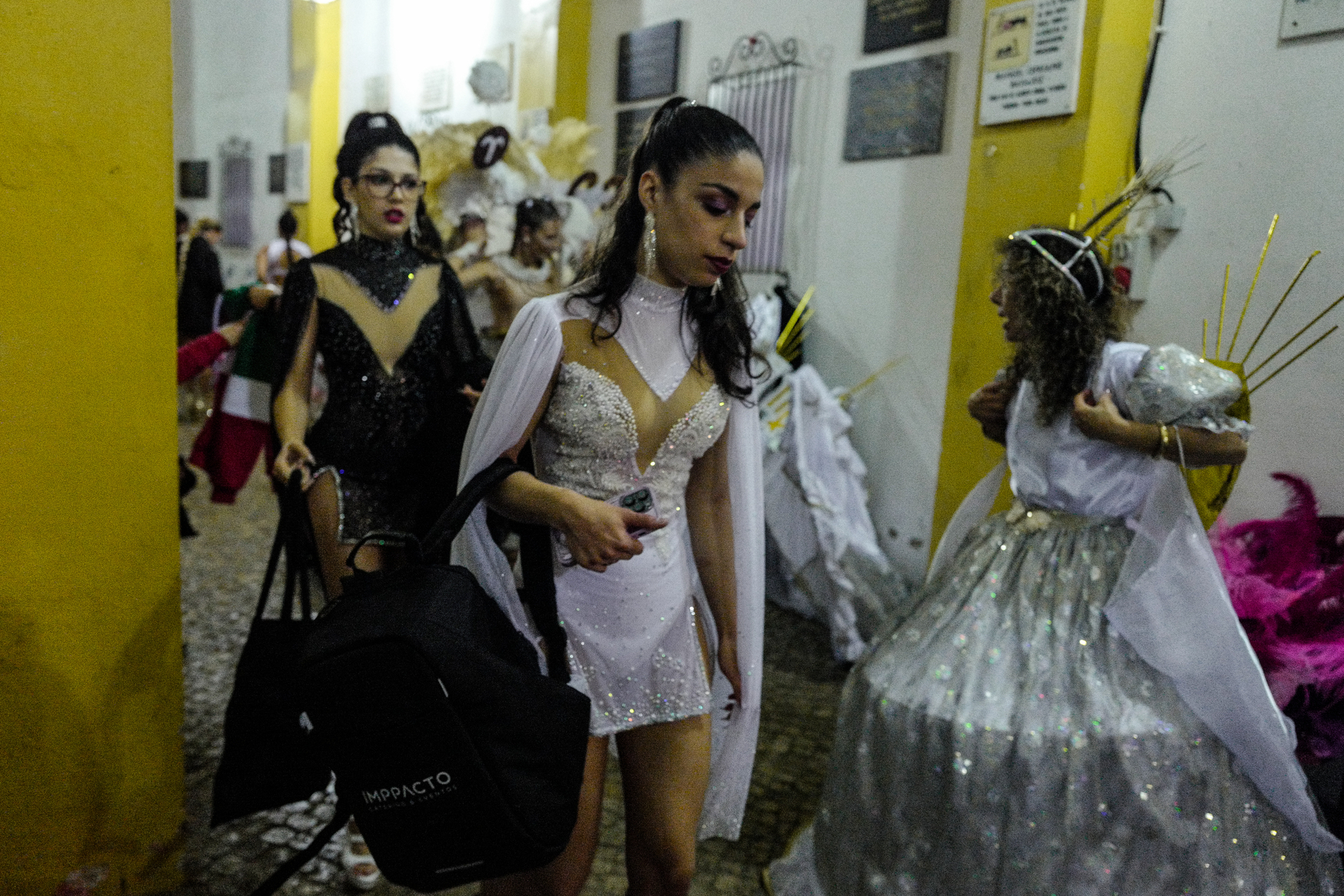 Individuals in elaborate costumes, possibly dancers or performers, are inside a building with yellow walls, preparing for an event. The focus is on a woman wearing a white dress adorned with rhinestones, holding a black shoulder bag.