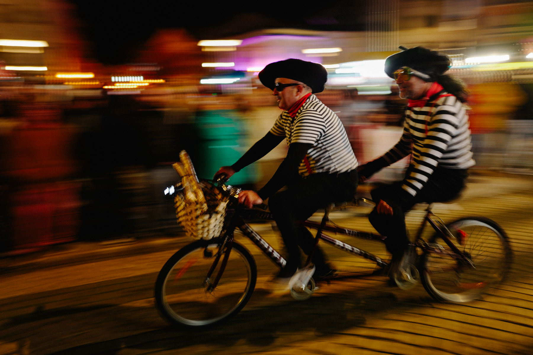 Two people in striped shirts and berets riding a tandem bicycle at night, with motion blur emphasizing speed and urban nightlife in the background.