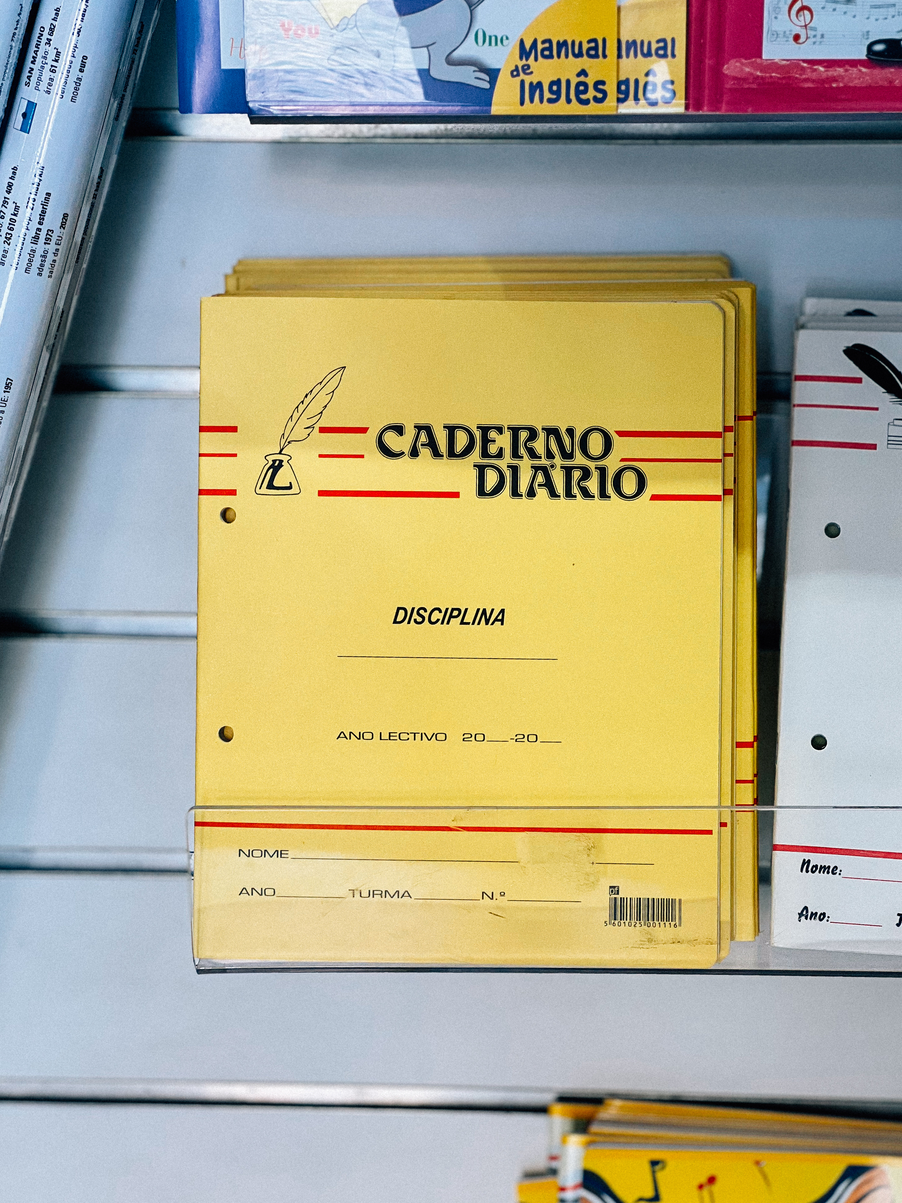 A stack of yellow school notebooks labeled &ldquo;Caderno Diario&rdquo; with space for discipline, school year, name, class, and number on a bookshelf.