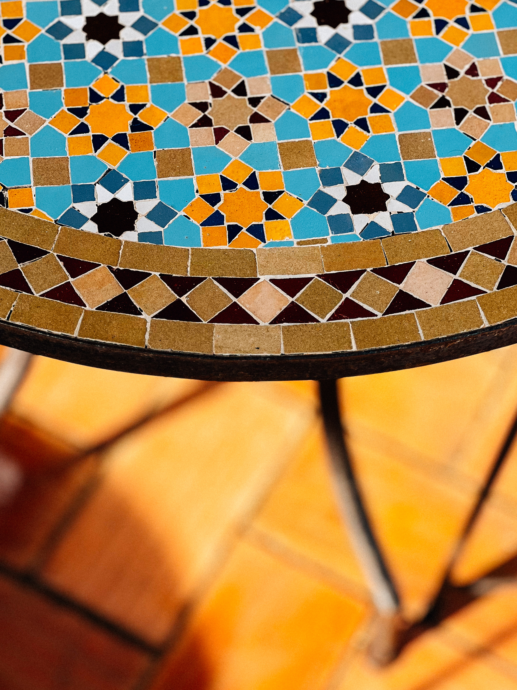 A close-up of a colorful mosaic tile tabletop with a pattern of geometric shapes in shades of blue, orange, and white, set against a blurry background of terracotta floor tiles.