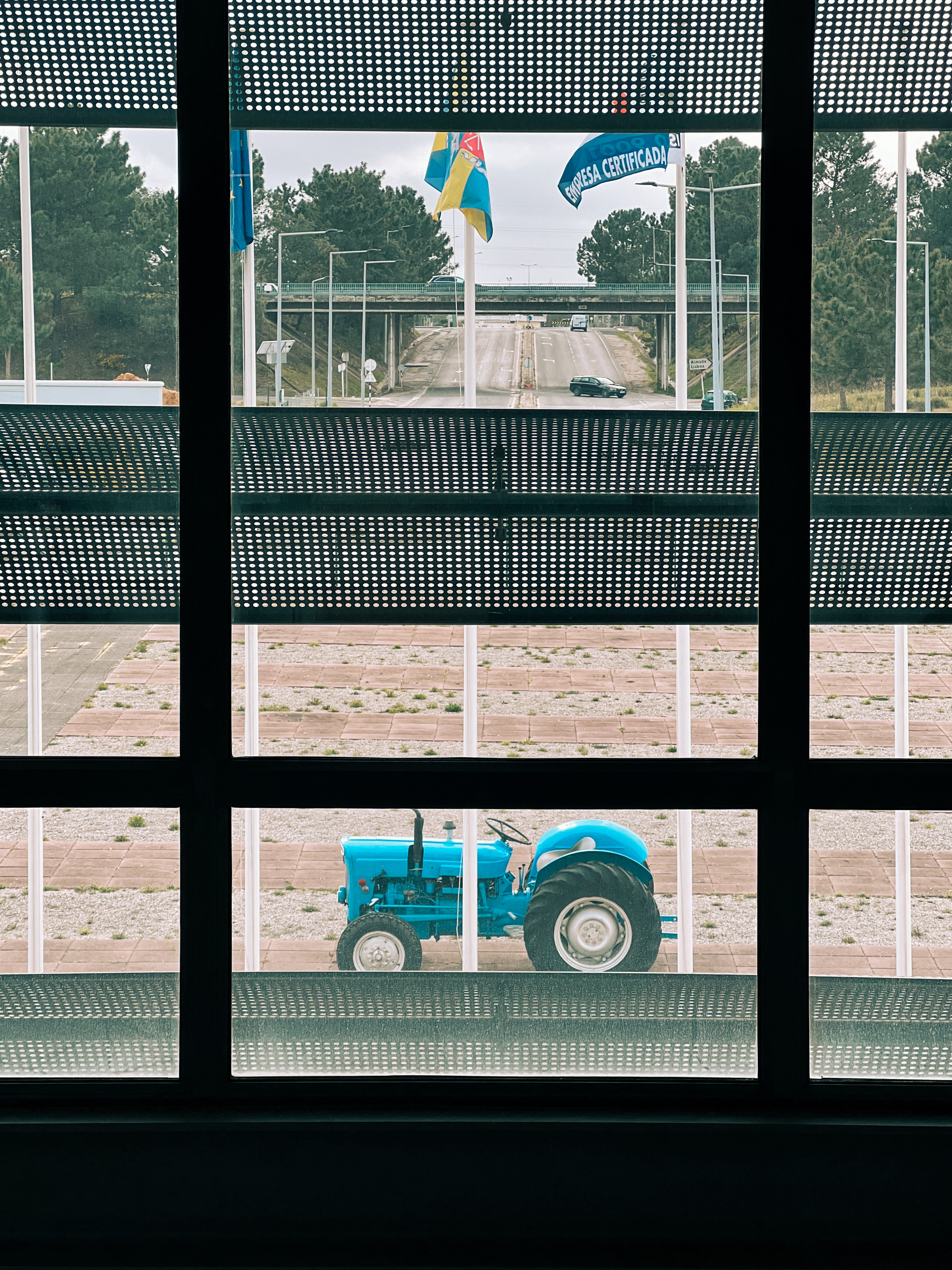 A view through a window with patterned shading revealing a blue tractor parked outside, with a highway in the background flanked by greenery and two flags fluttering in the wind.
