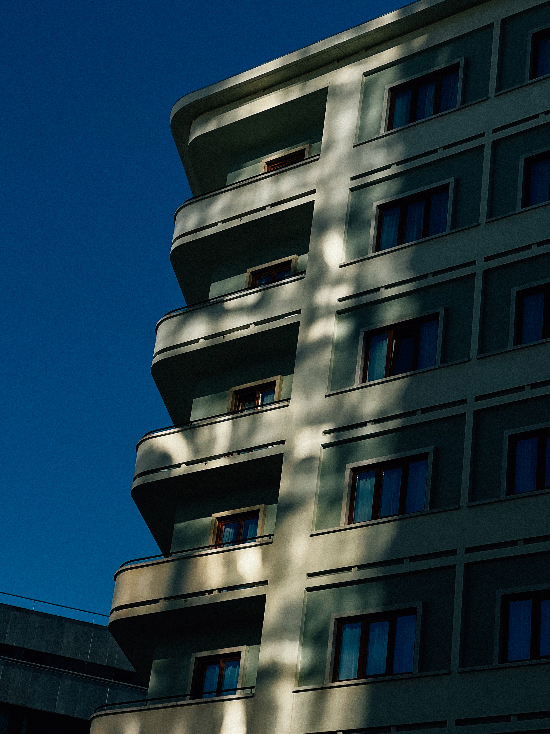 A modern building with curved balconies, featuring shadows and sunlight on its facade under a clear blue sky.