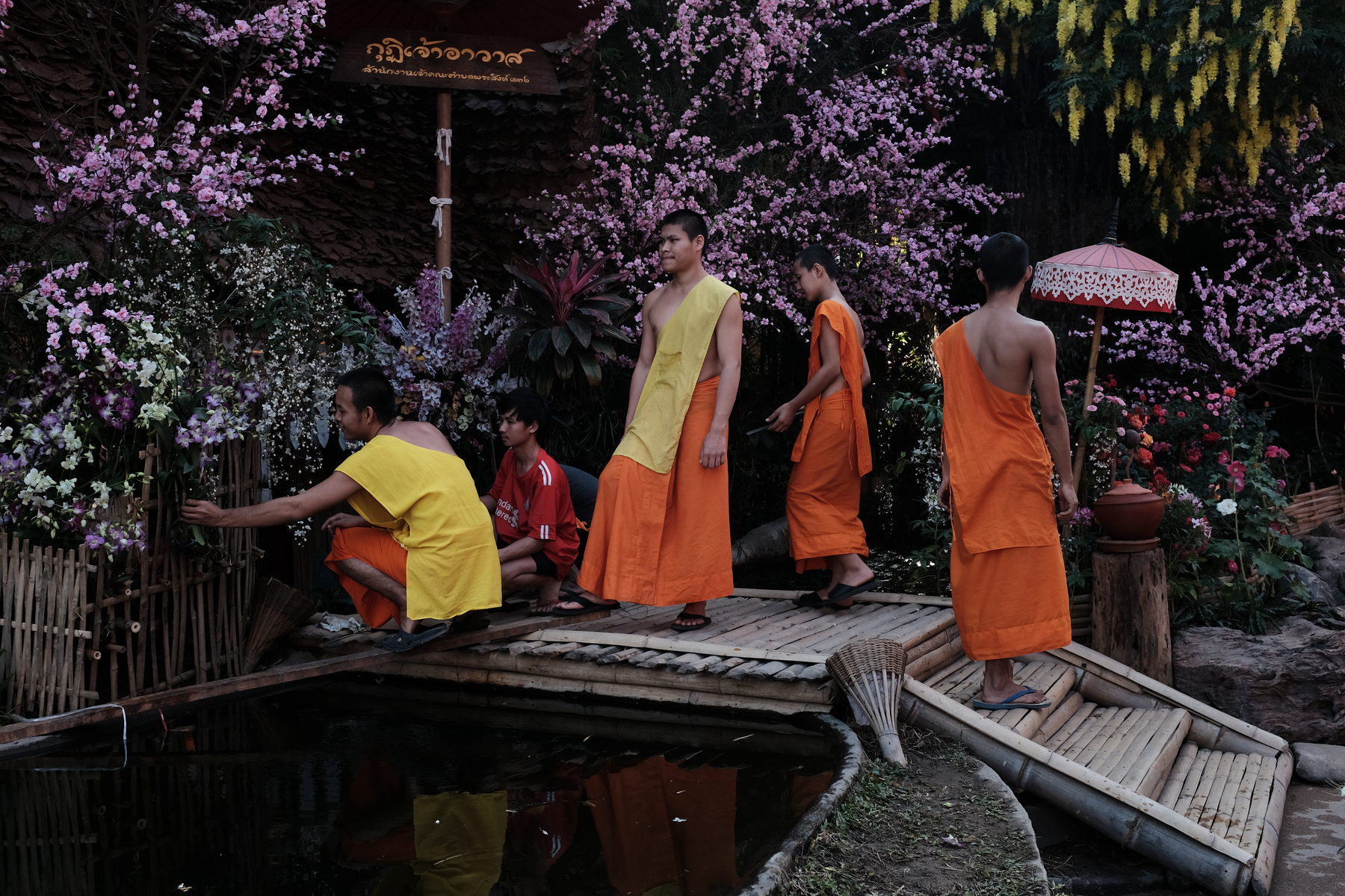 Individuals in traditional orange robes, likely Buddhist monks, walking across a bamboo bridge with a background of colorful flowers.