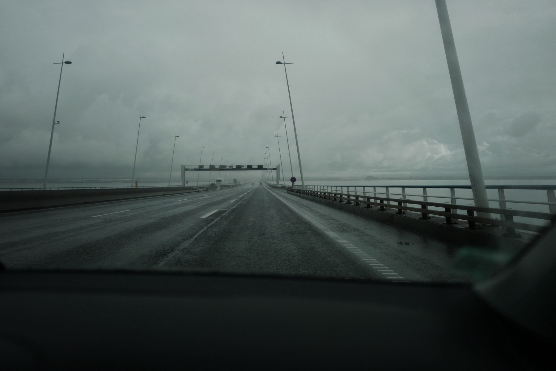 View from inside a car approaching a bridge during rainy weather, with overcast skies and wet road surface. Street lights line the sides of the bridge.