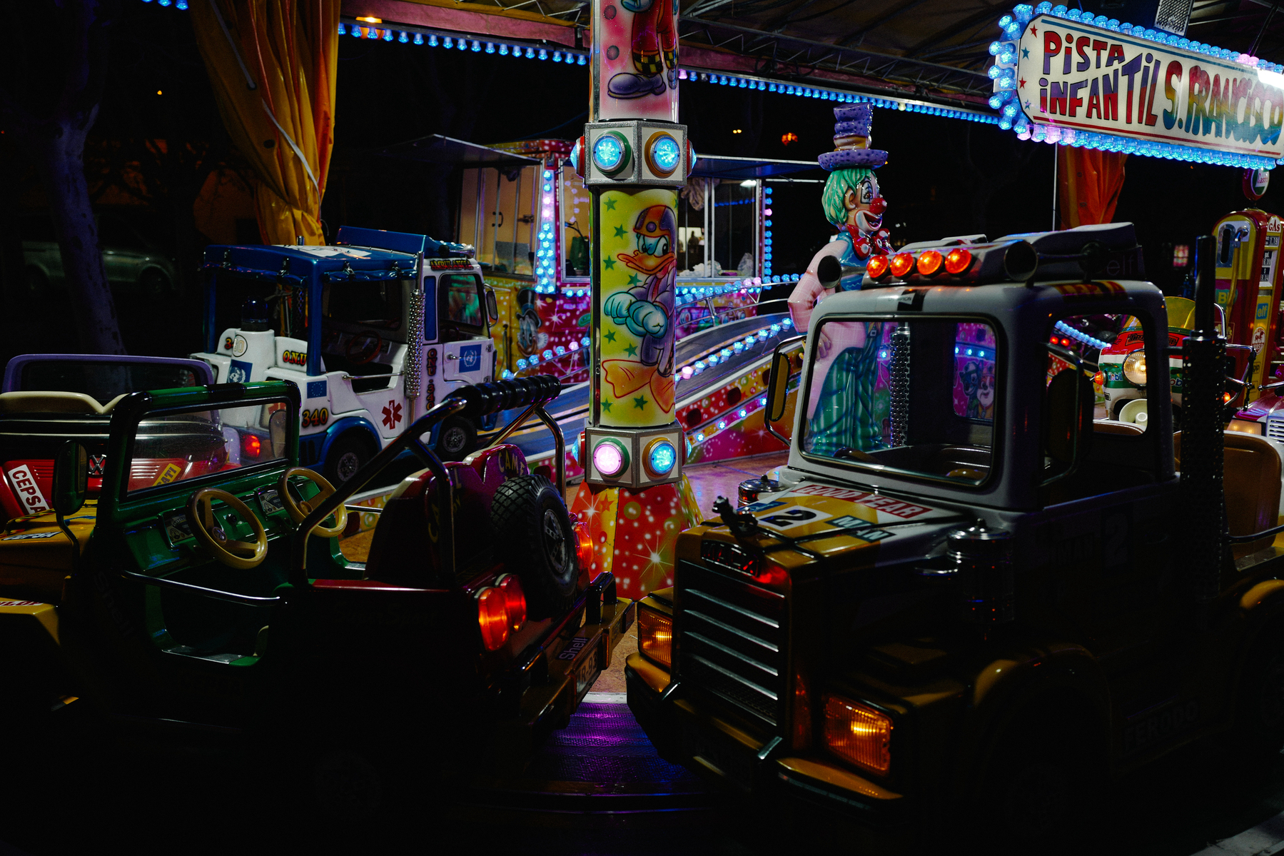 Amusement park ride featuring colorful kiddie trucks adorned with lights on a vibrant track, decorated with circus-themed artwork, at night.