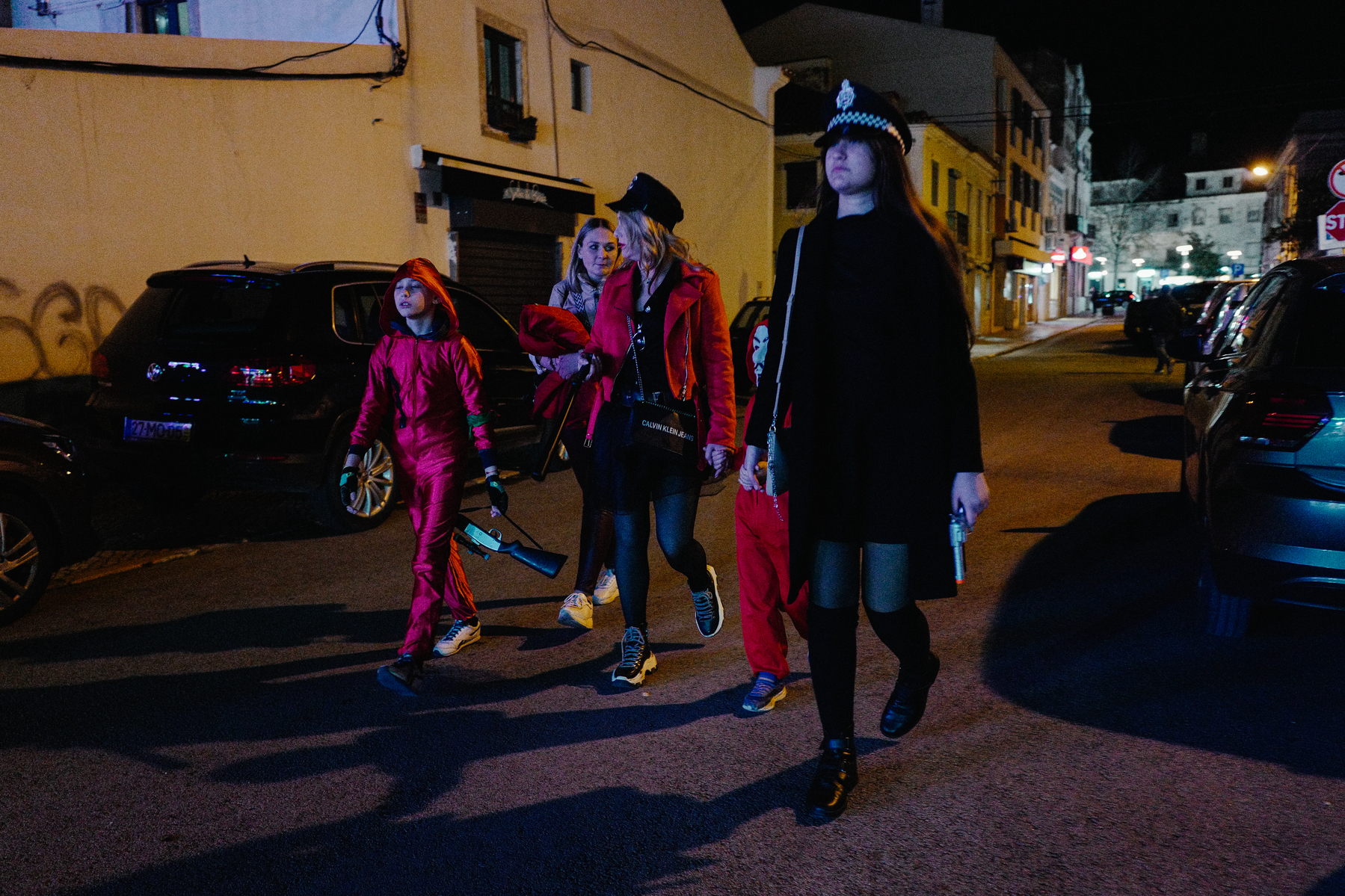 A group of individuals walking at night on a city street. One person is wearing a vibrant pink suit, another is in a red jacket with a graphic print, and a third is dressed in a black coat with a police hat.