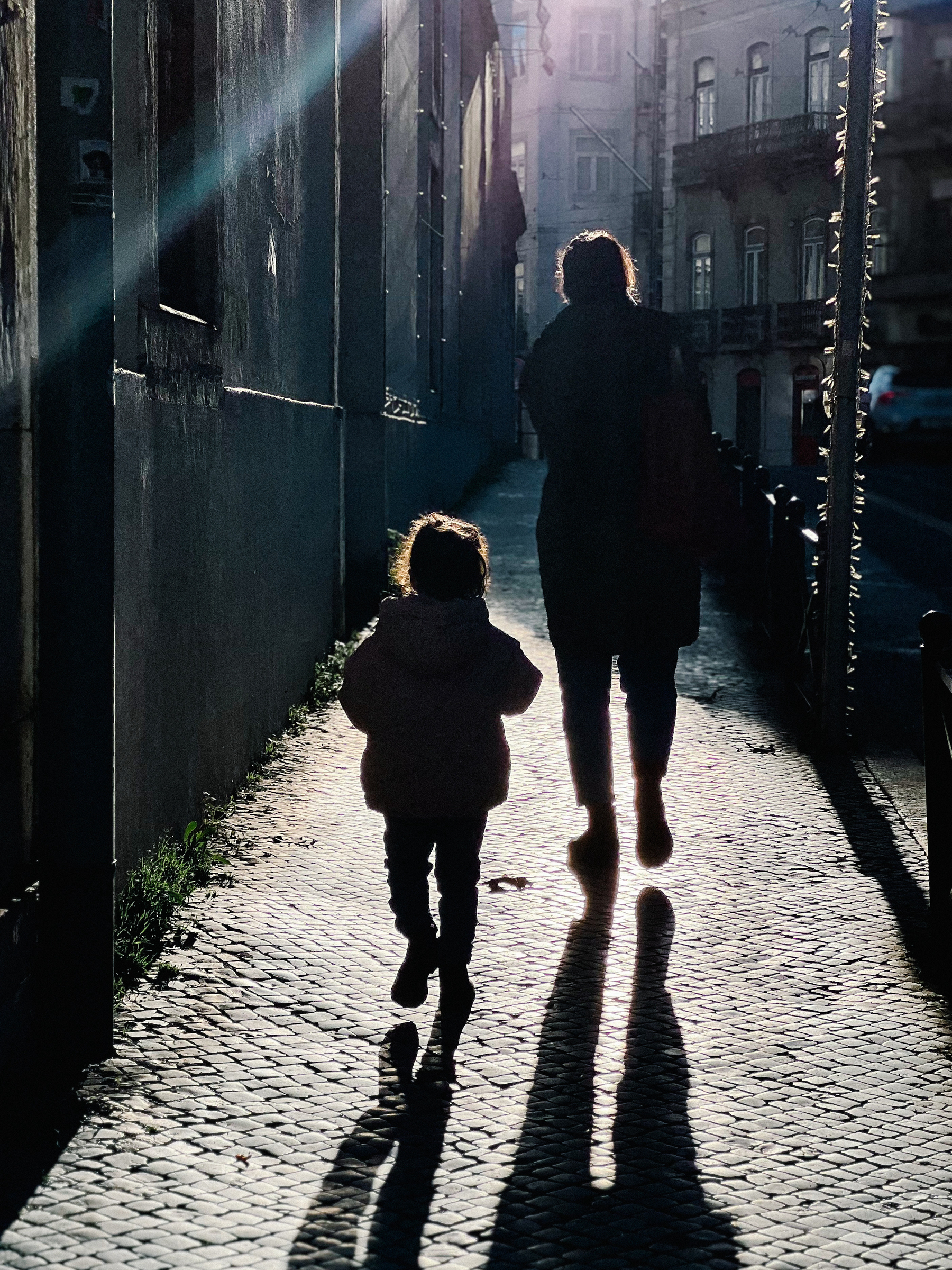 A silhouette of an adult and a child walking down a cobblestone street, with long shadows stretching out in front of them due to the low-angle sunlight.