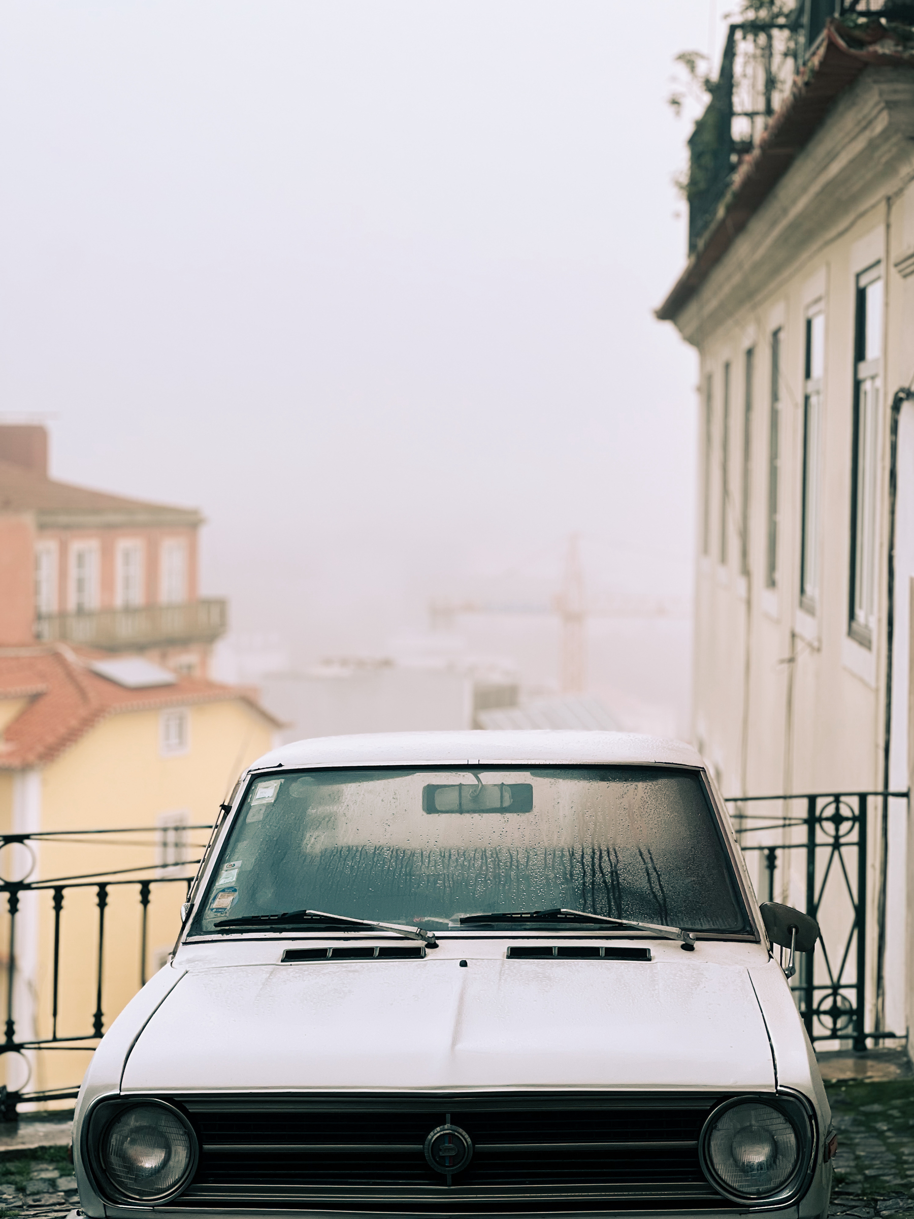 A vintage white car parked on a cobblestone street next to a building with railings, with a foggy cityscape in the background.