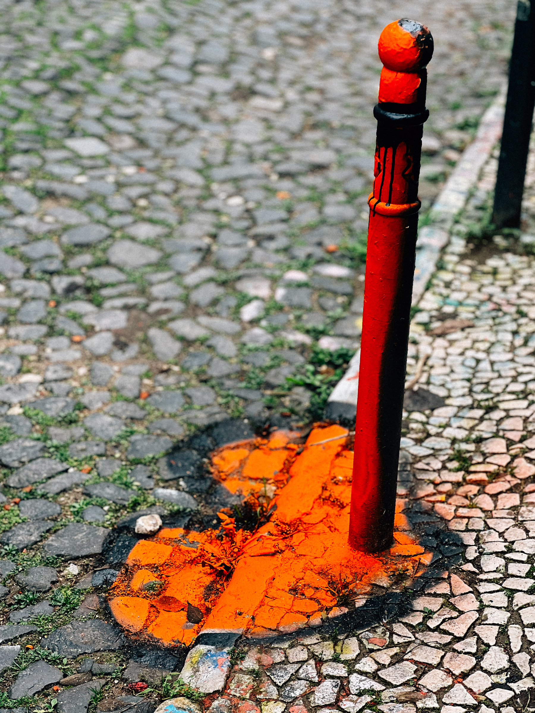 An orange painted metal bollard on a cobblestone street with orange paint splashed at its base.