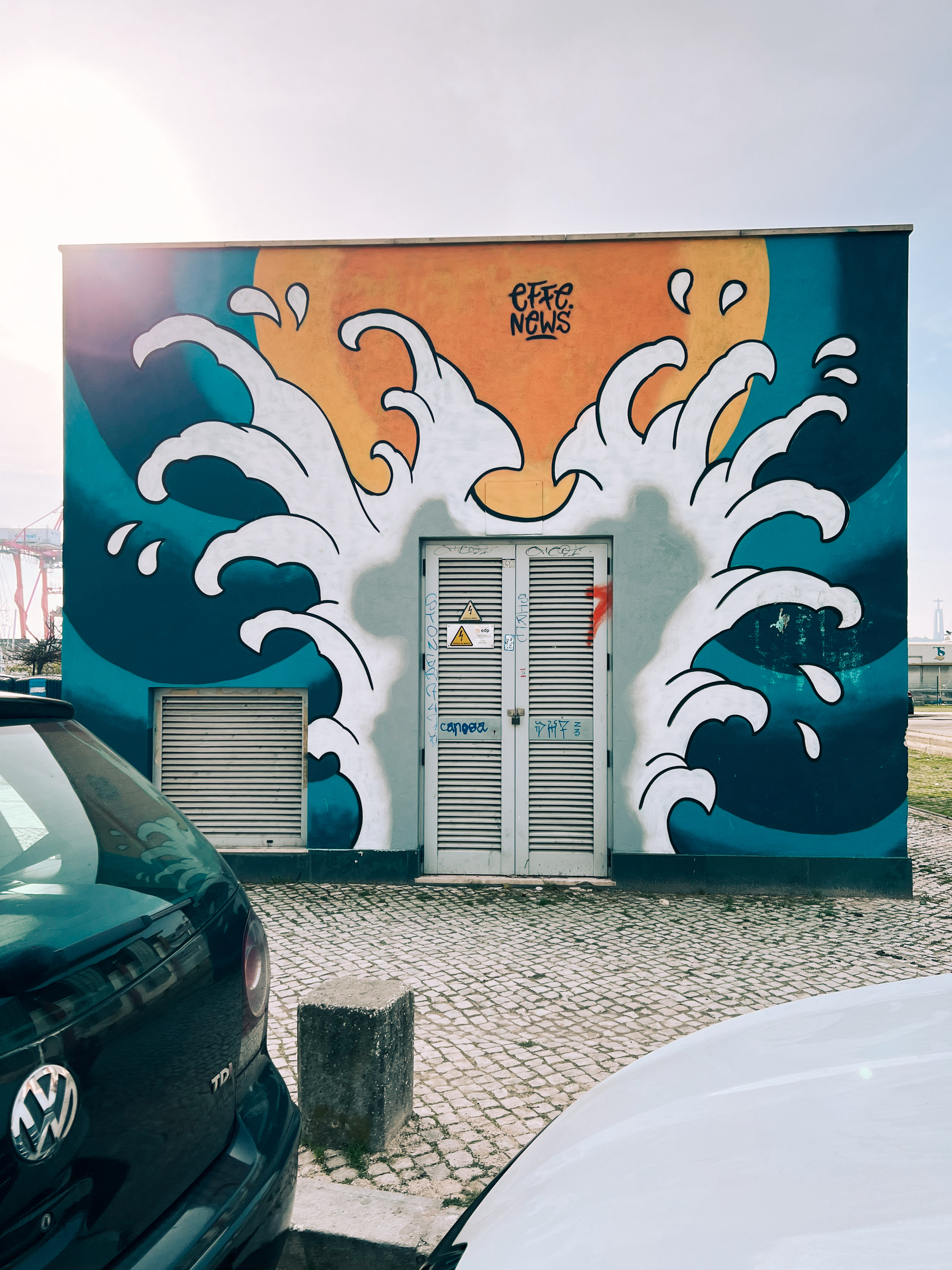 A colorful mural with wave designs on a building wall, featuring a blue and white color scheme with orange accents. The wall has two metal doors with graffiti tags, and two cars are partially visible in the foreground.