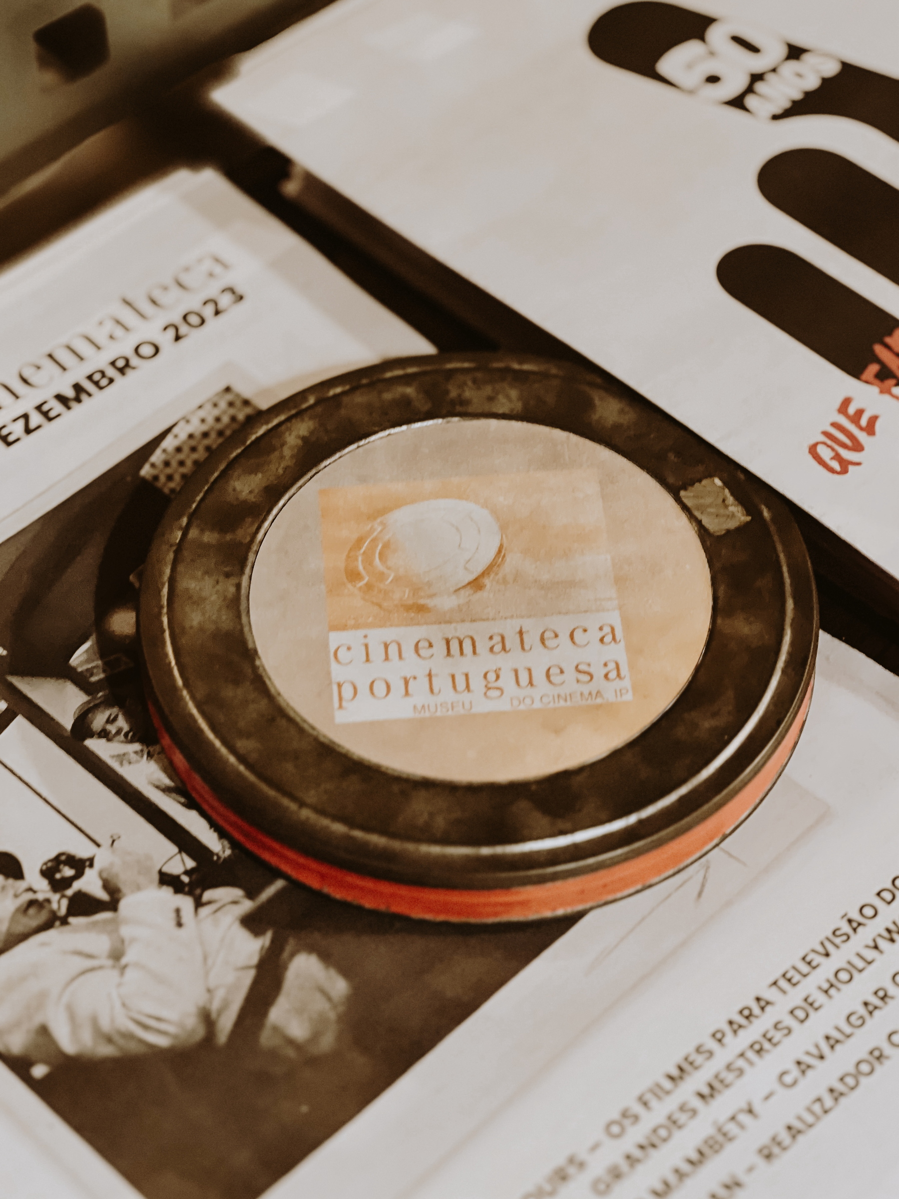 An old film canister on top of brochures or flyers, with text related to the &ldquo;Cinemateca Portuguesa&rdquo; (Portuguese Film Archive) and cinema-related content visible.