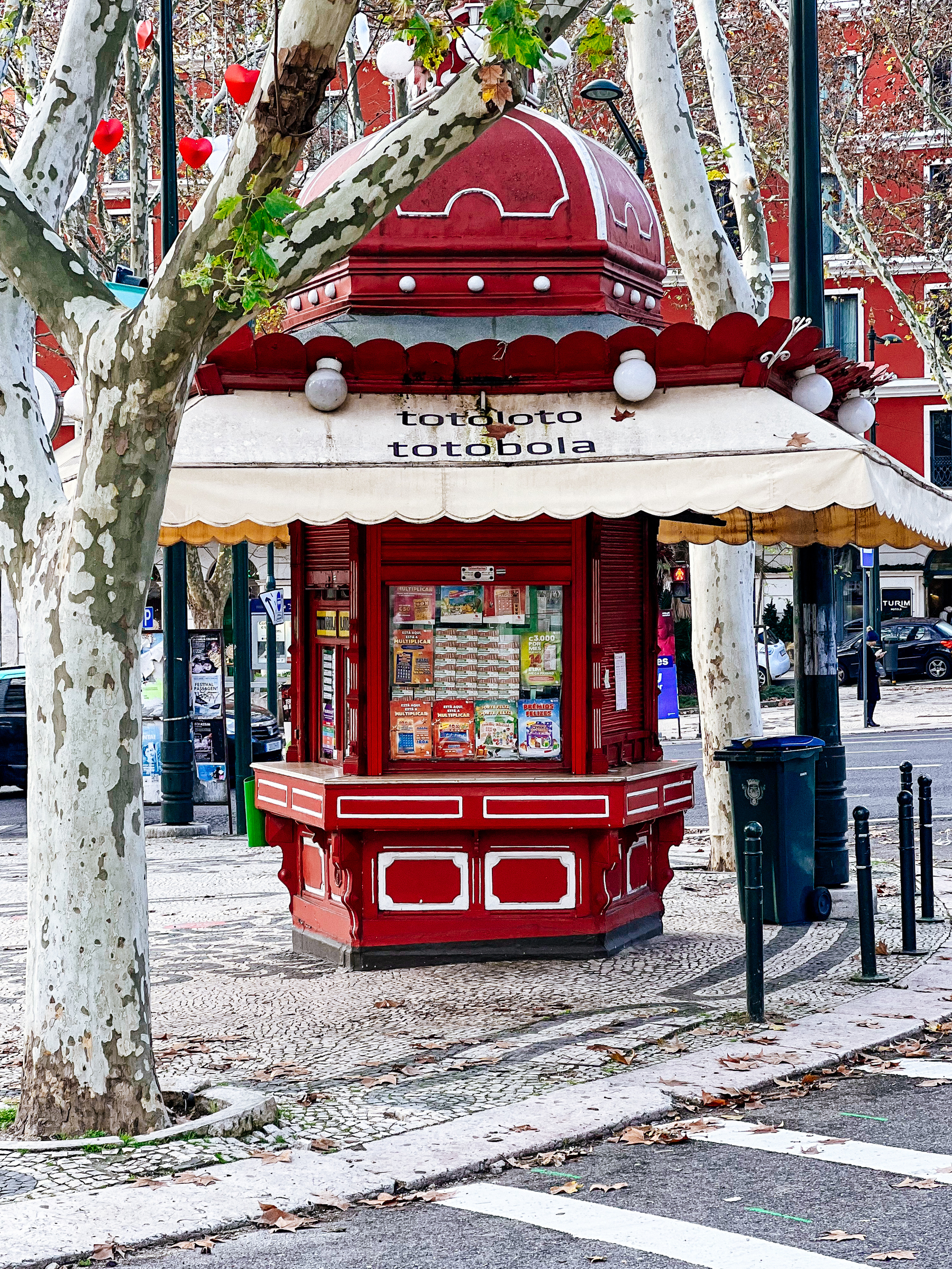 A vibrant red kiosk with &ldquo;TOTOLOTO TOTOBOLA&rdquo; lettering, nestled between two trees with mottled bark, on a cobblestone sidewalk. The kiosk has a dome-like top.
