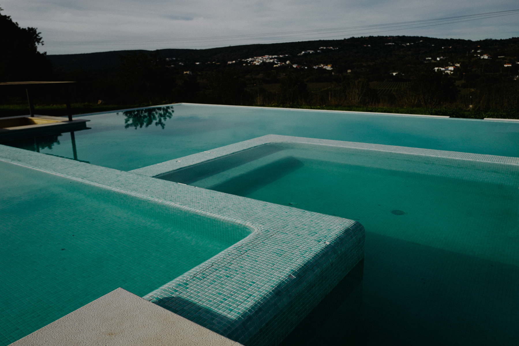 An outdoor swimming pool with clear blue water bordered by light poolside decking. The pool features a built-in step or bench, and is set against a backdrop of scenic green hills.