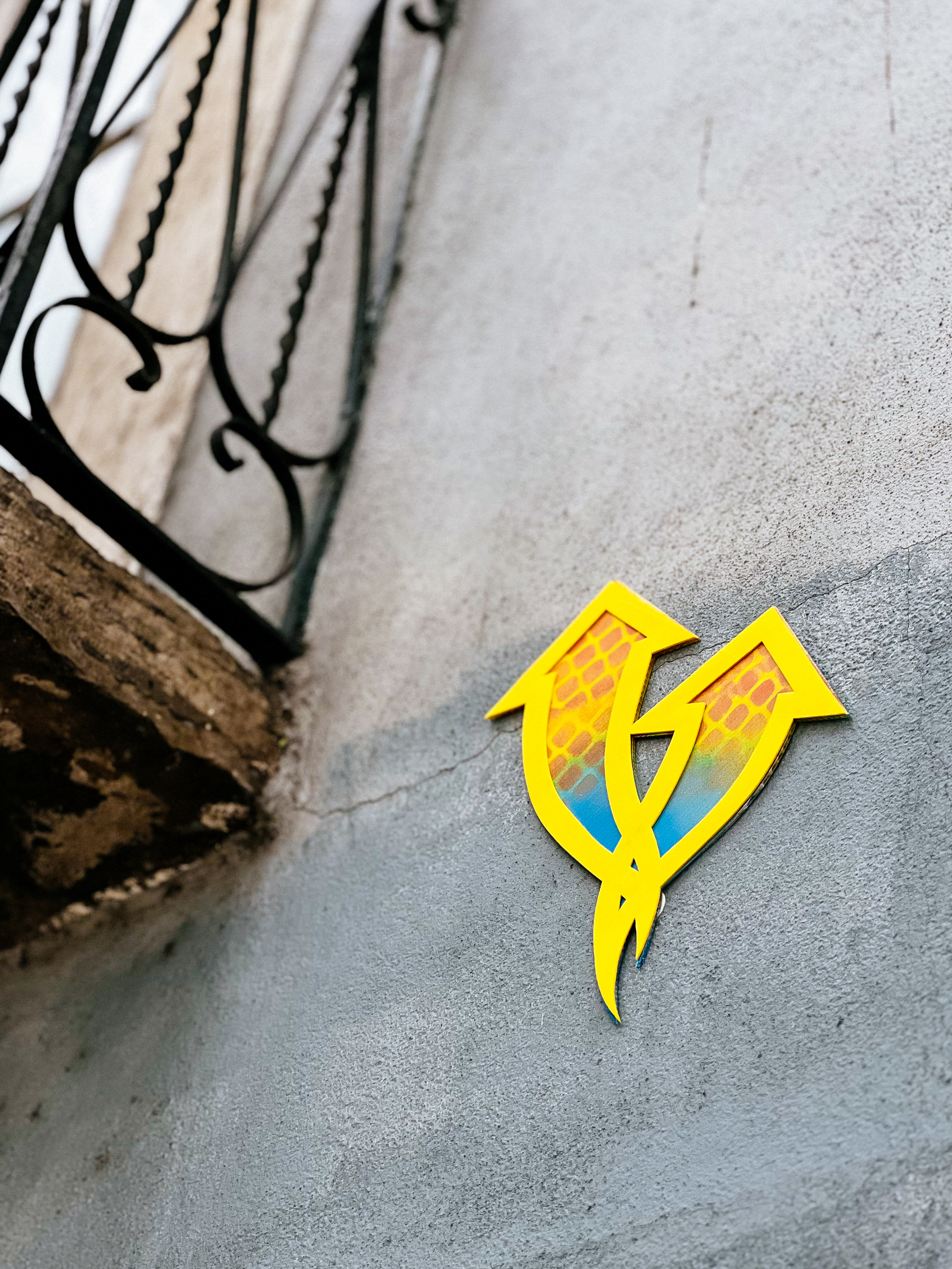 A yellow street art piece, a couple of intertwined arrows, attached to a gray concrete wall next to a wrought iron balcony.