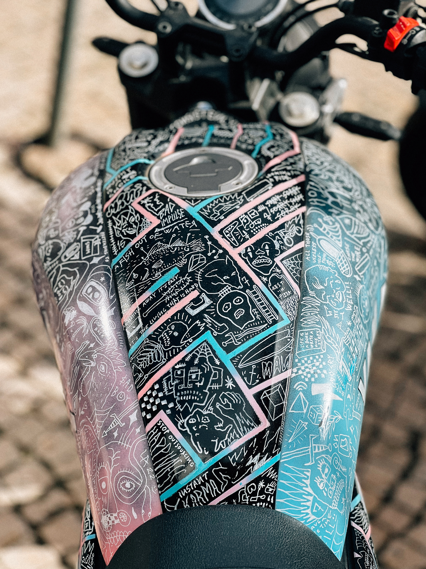 A motorcycle tank covered with colorful, detailed graffiti-style artwork, in focus against a blurry background of motorcycle parts.