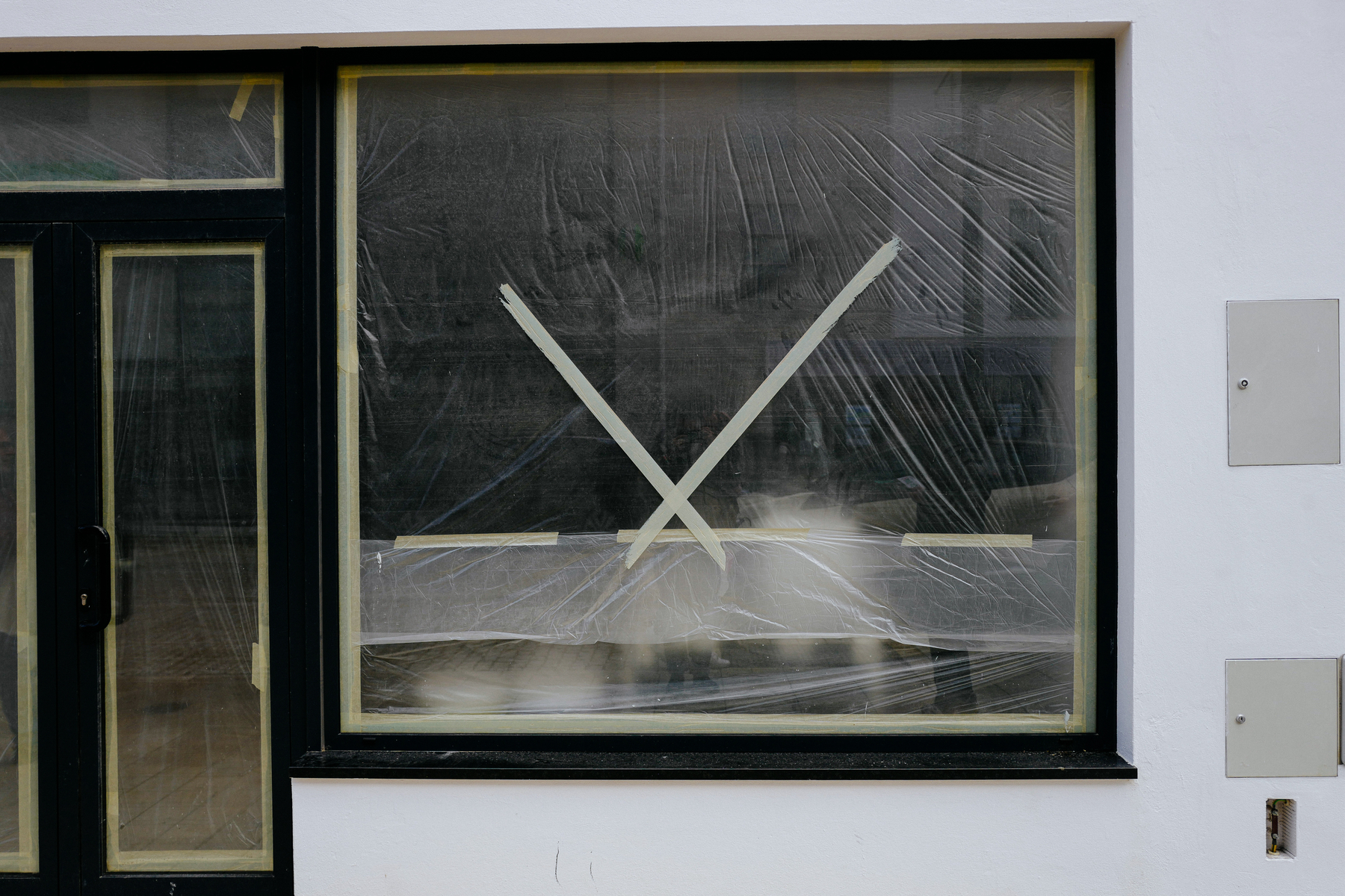 A taped-up window with protective plastic covering, suggesting construction or maintenance, on a building&rsquo;s exterior wall beside a closed door and wall-mounted utility covers.