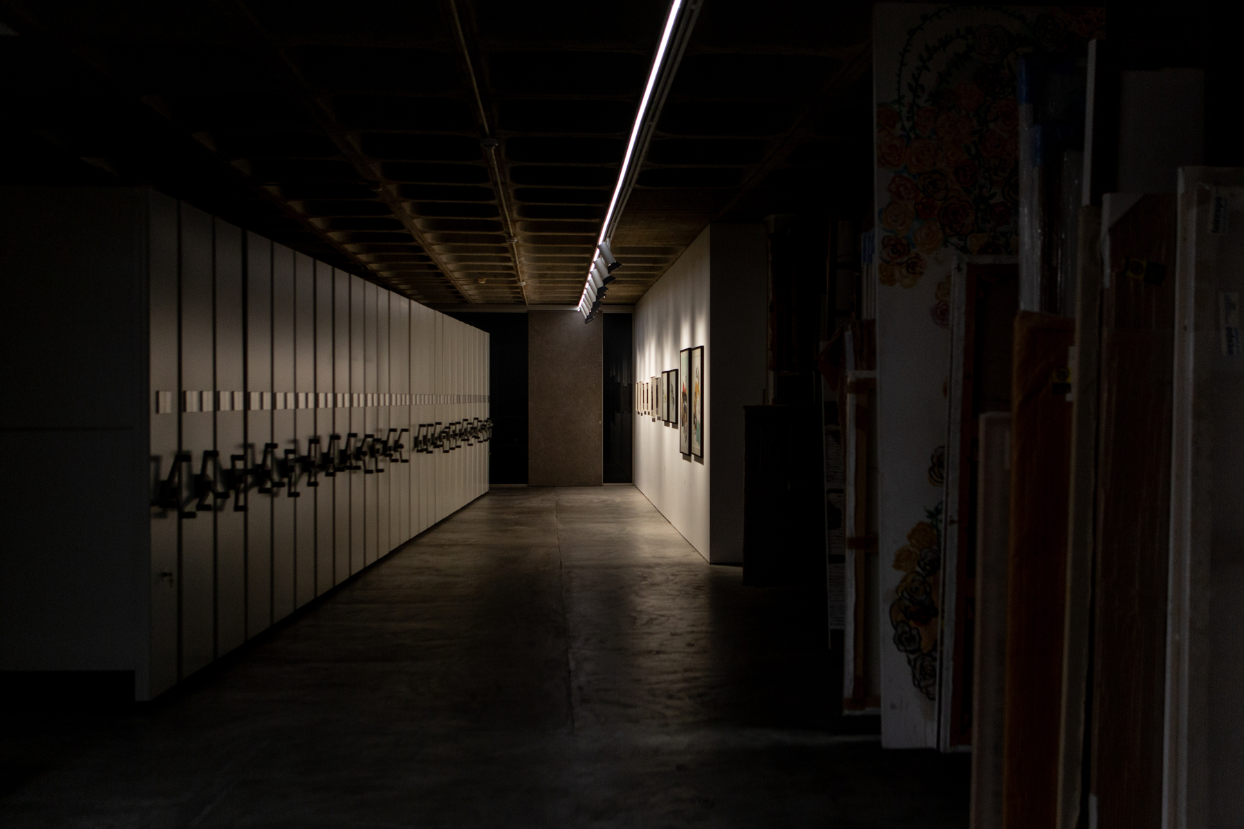 A dimly lit corridor flanked by lockers on the left and framed artworks on the right, with overhead fluorescent lighting and stacked canvases to the far right.