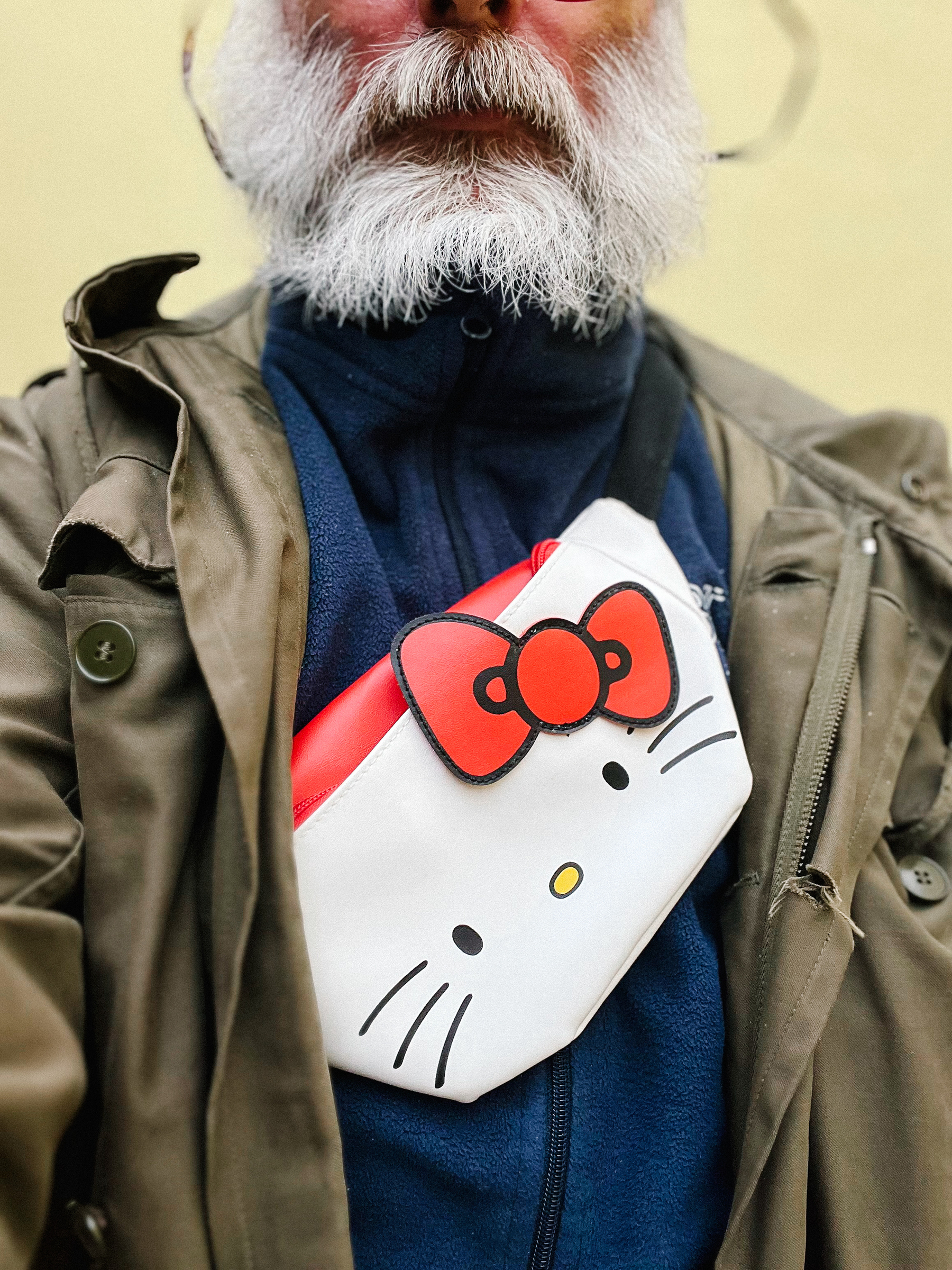 A person with a white beard wearing a bumbag with a Hello Kitty design, a blue fleece jacket, and an olive green jacket.
