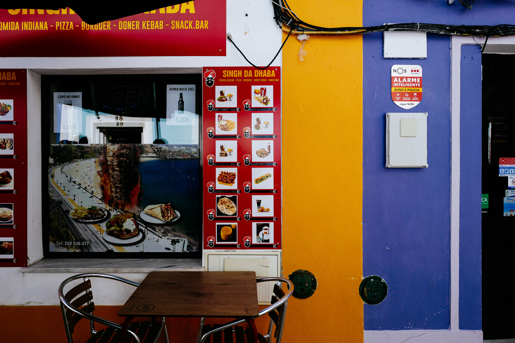 A colorful street view of a restaurant exterior with a red sign that reads &ldquo;COMIDA INDIANA - PIZZA - BURGUER - DONER KEBAB - SNACK.BAR.&rdquo; A photo menu is displayed beside the entrance,