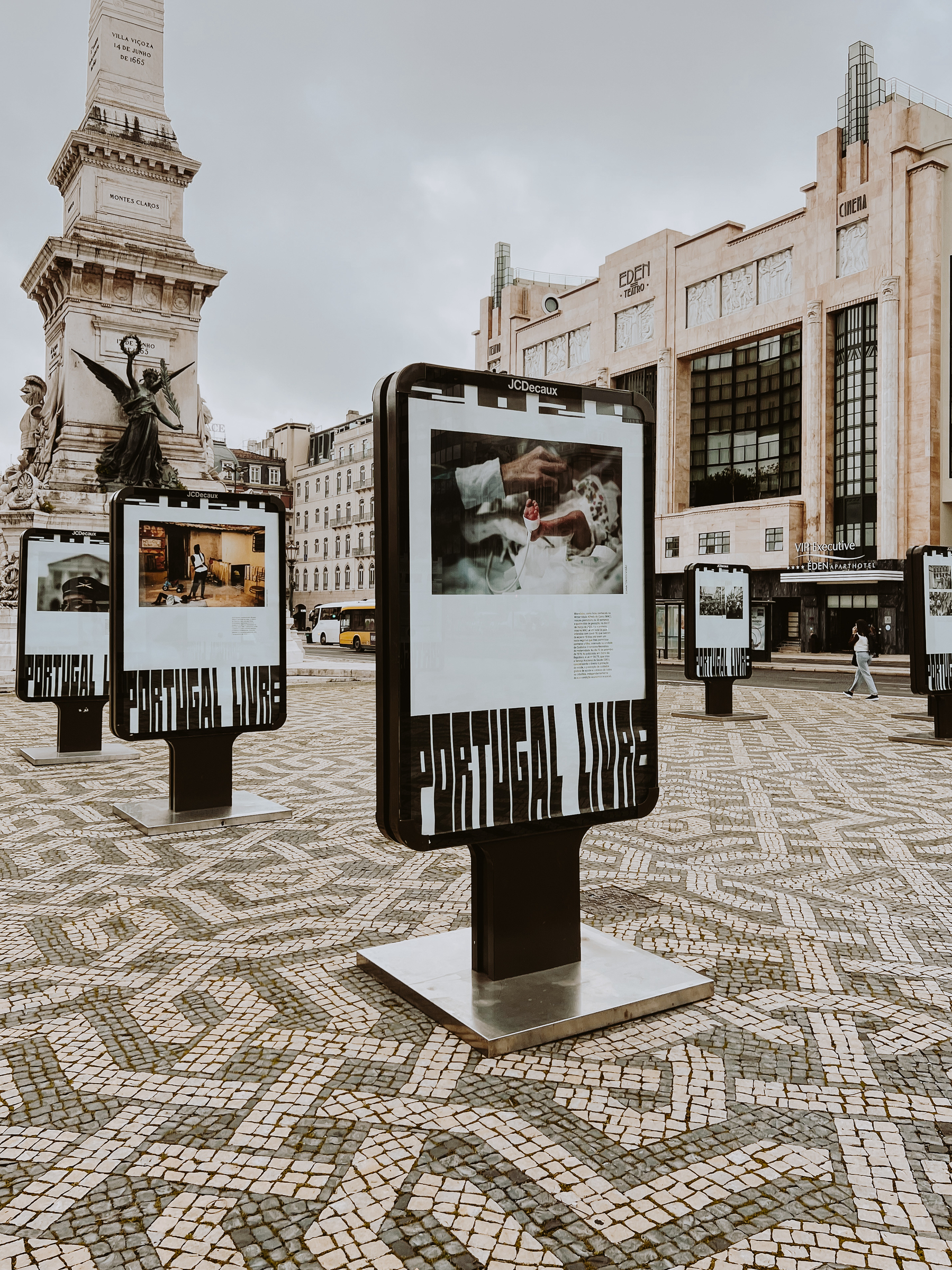 Outdoor exhibition displays with photographs on a public square with cobblestone pavement, and a classical-style monument in the background. Text on the photographs reads &ldquo;PORTUGAL LIVRE.&quot;