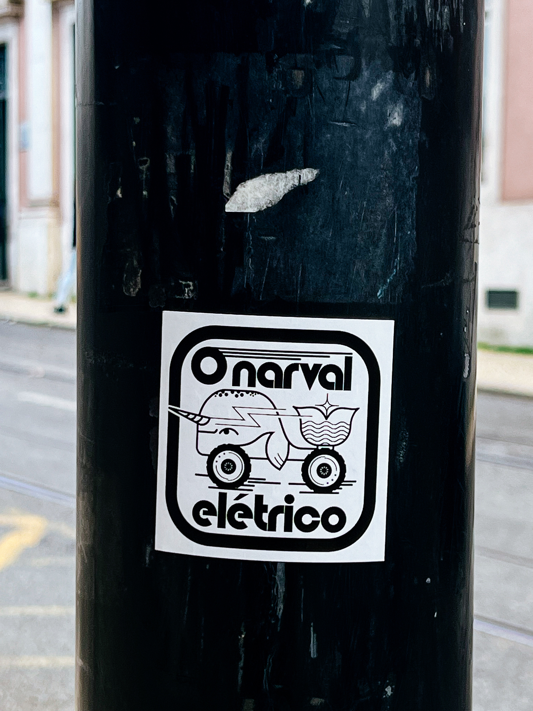 A sticker on a dark pole depicts a cartoon narwhal on wheels with the text &ldquo;O narval eléctrico&rdquo;.