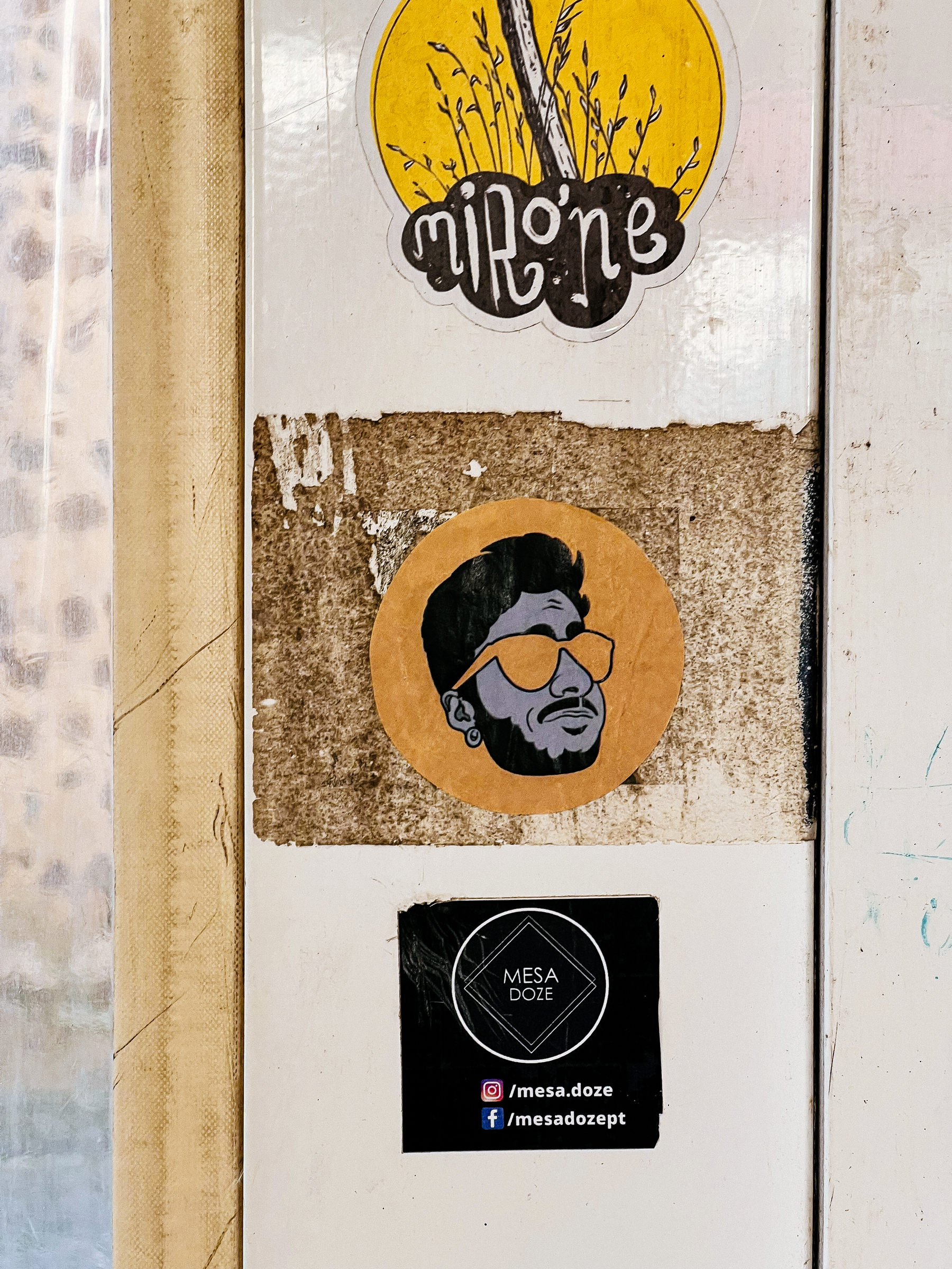 Stickers on a worn surface show a portrait and logos. They depict a bearded man wearing glasses and two brand names with social media handles.