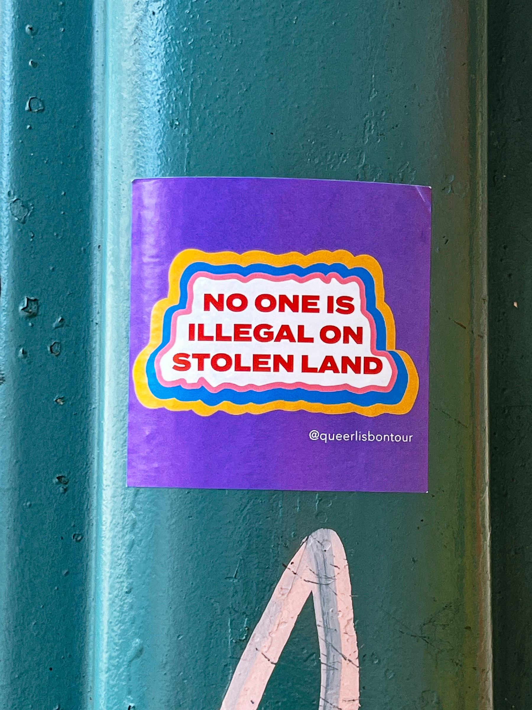 A purple sticker with yellow and red borders displays the text &ldquo;NO ONE IS ILLEGAL ON STOLEN LAND @queerlisbontour&rdquo; affixed to a green metal pole.