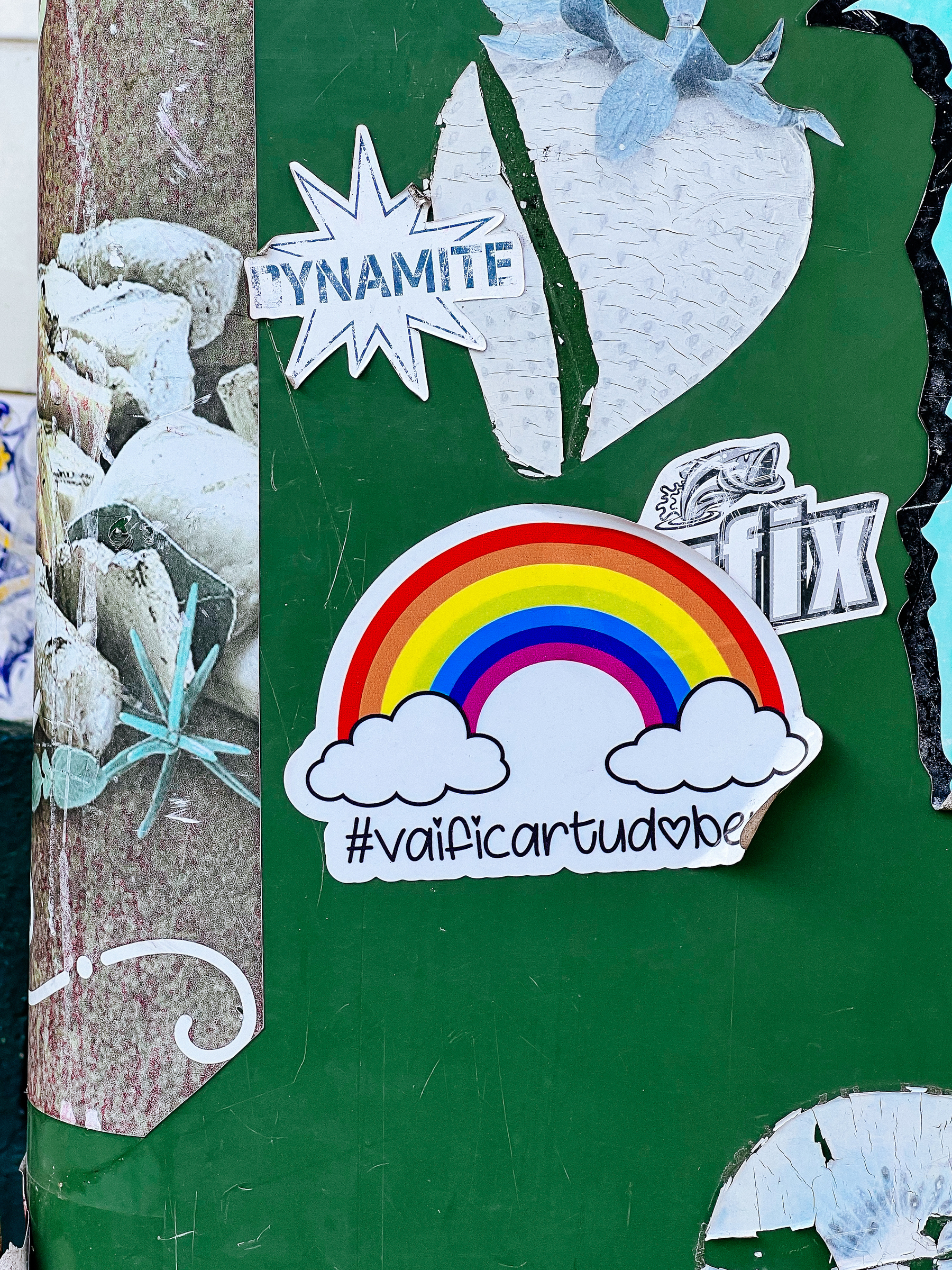 A rainbow sticker with text &ldquo;#vaificartudobem&rdquo; affixed to a weathered green surface surrounded by other worn stickers and graffiti elements.