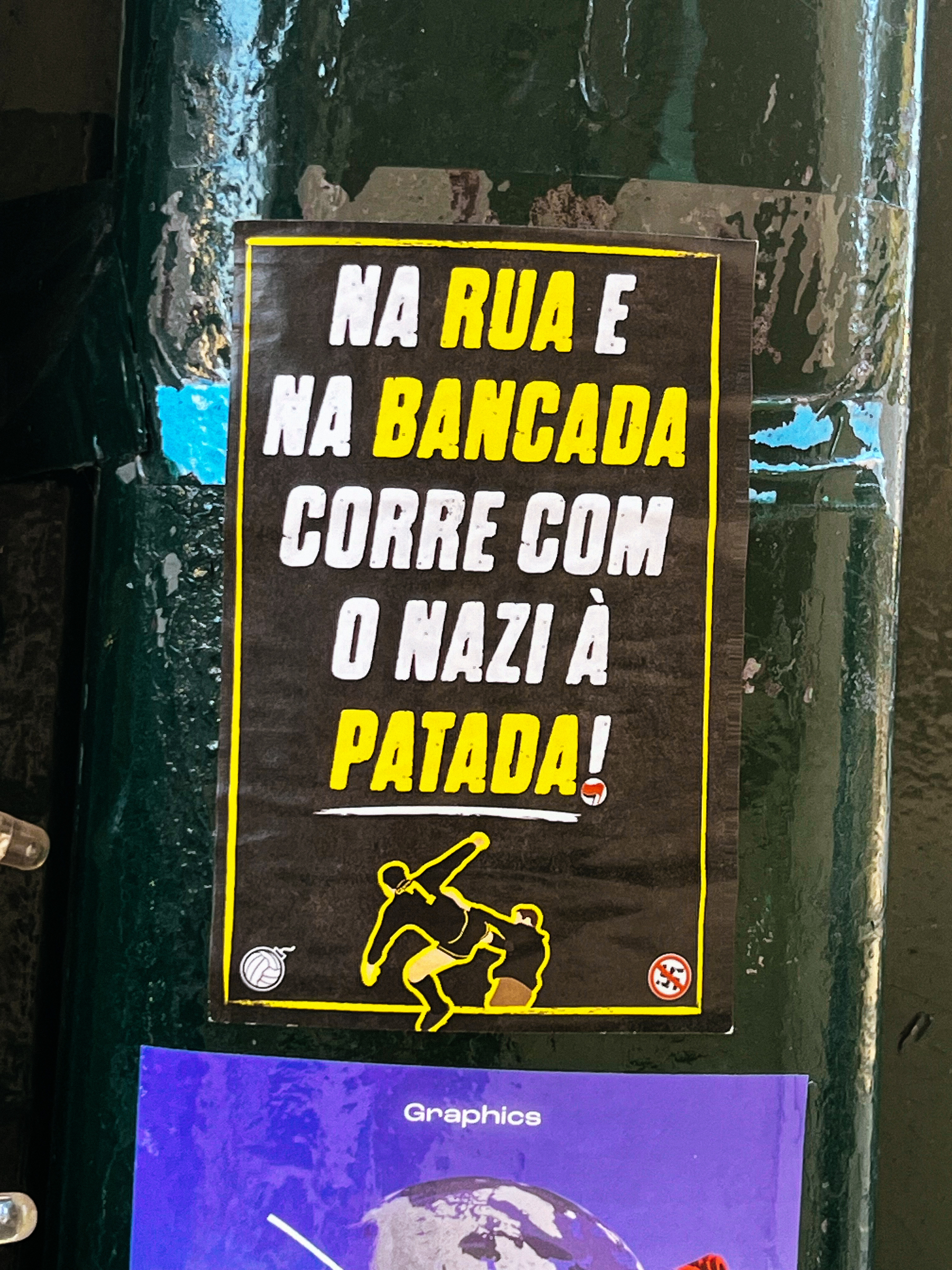 A yellow and black sticker on a pole depicts a figure kicking another figure, with text &ldquo;NA RUA E NA BANCADA CORRE COM O NAZI À PATADA!&rdquo; indicating an anti-fascist message.