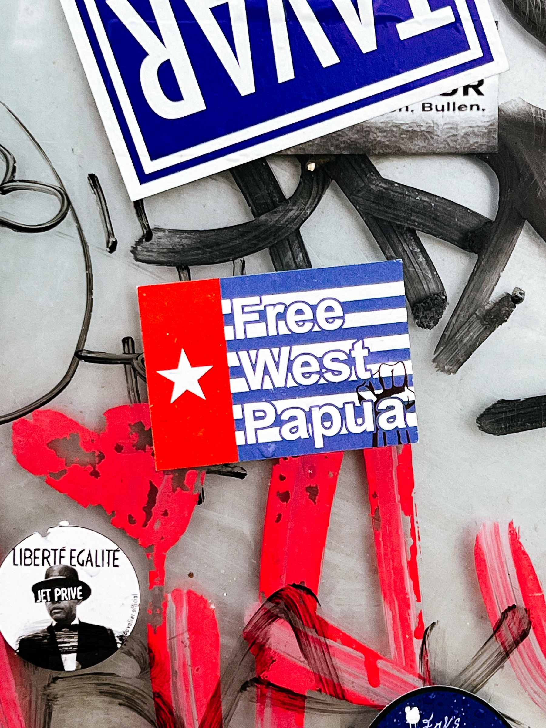 Stickers and graffiti overlay a surface, prominently featuring &ldquo;Free West Papua&rdquo; and &ldquo;LIBERTÉ ÉGALITÉ JET PRIVÉ,&rdquo; amidst abstract red and black paint marks.